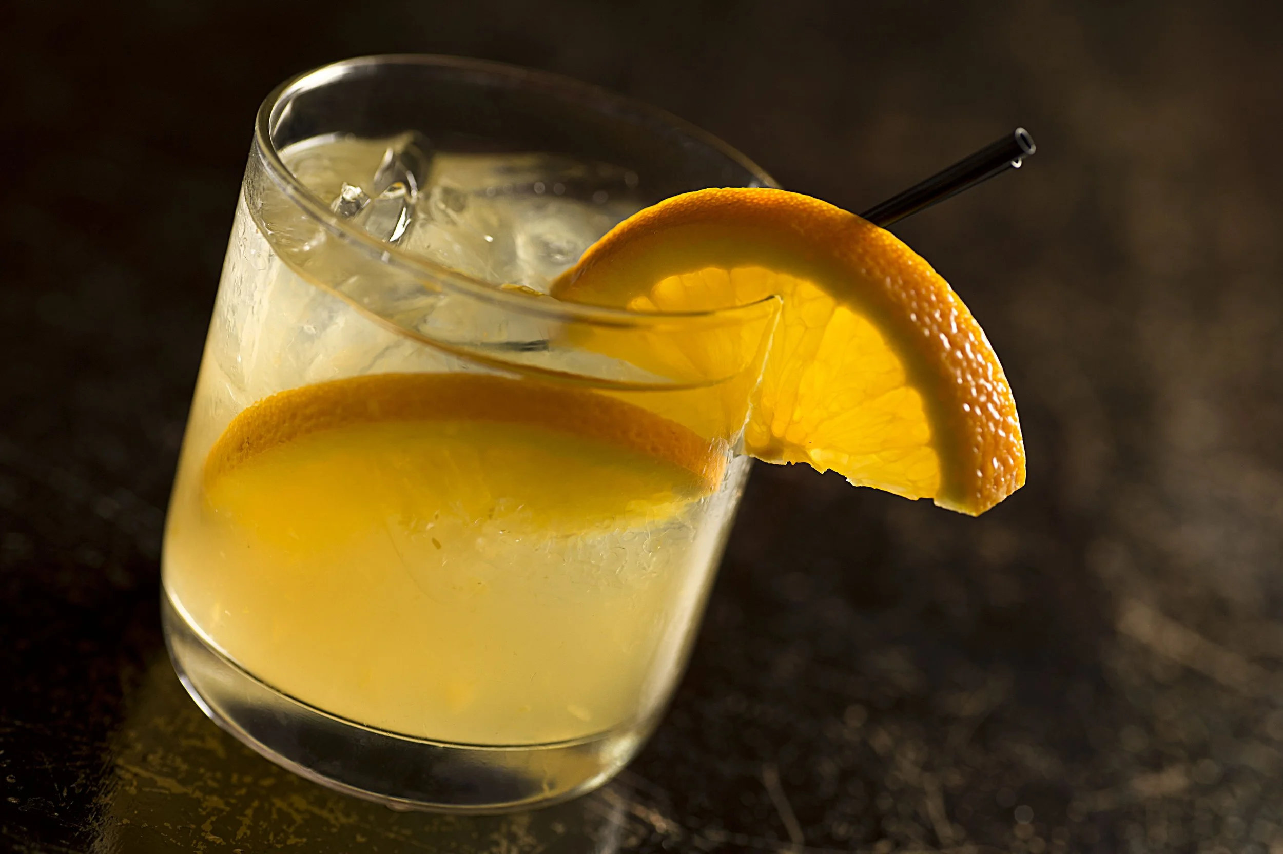 Close-up of a cocktail with ice, an orange slice garnish, and a straw in a glass.