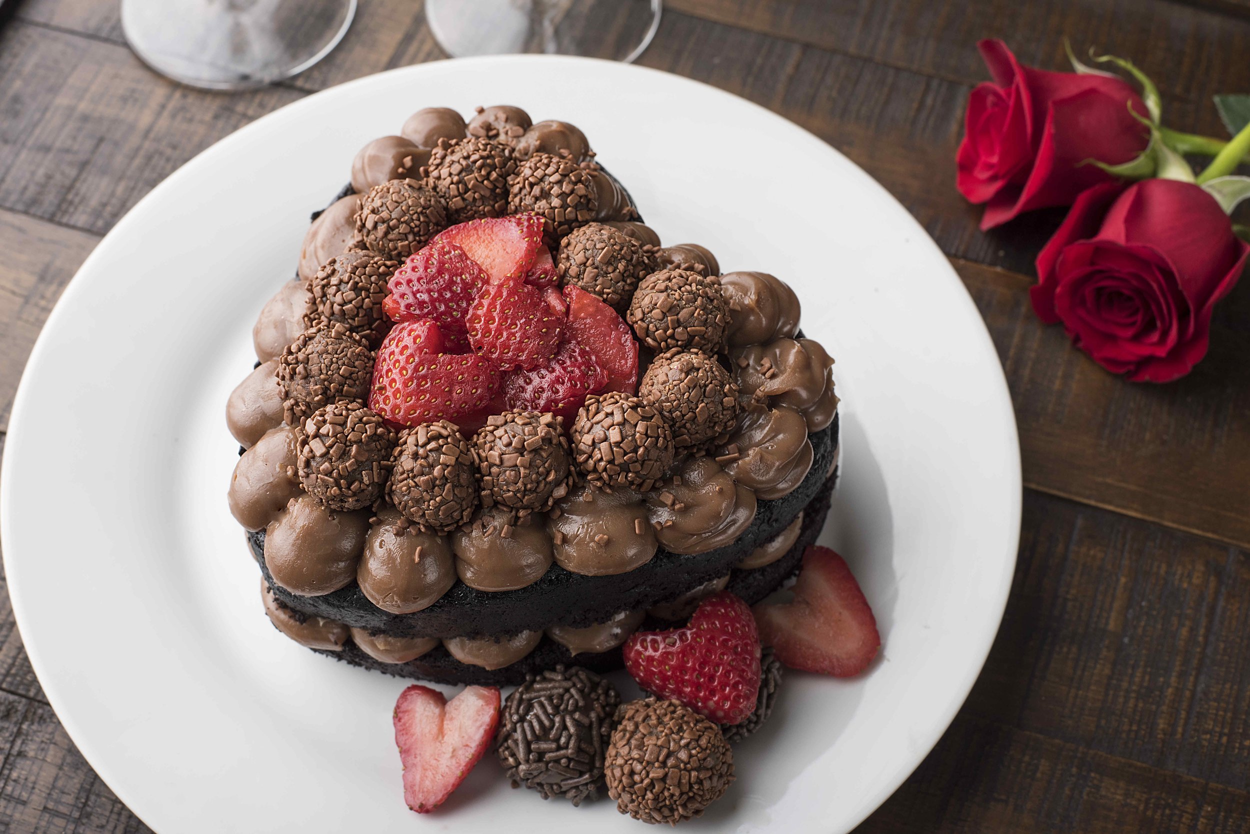 Heart-shaped chocolate cake with strawberries and chocolate truffles on a white plate, two red roses on the side.