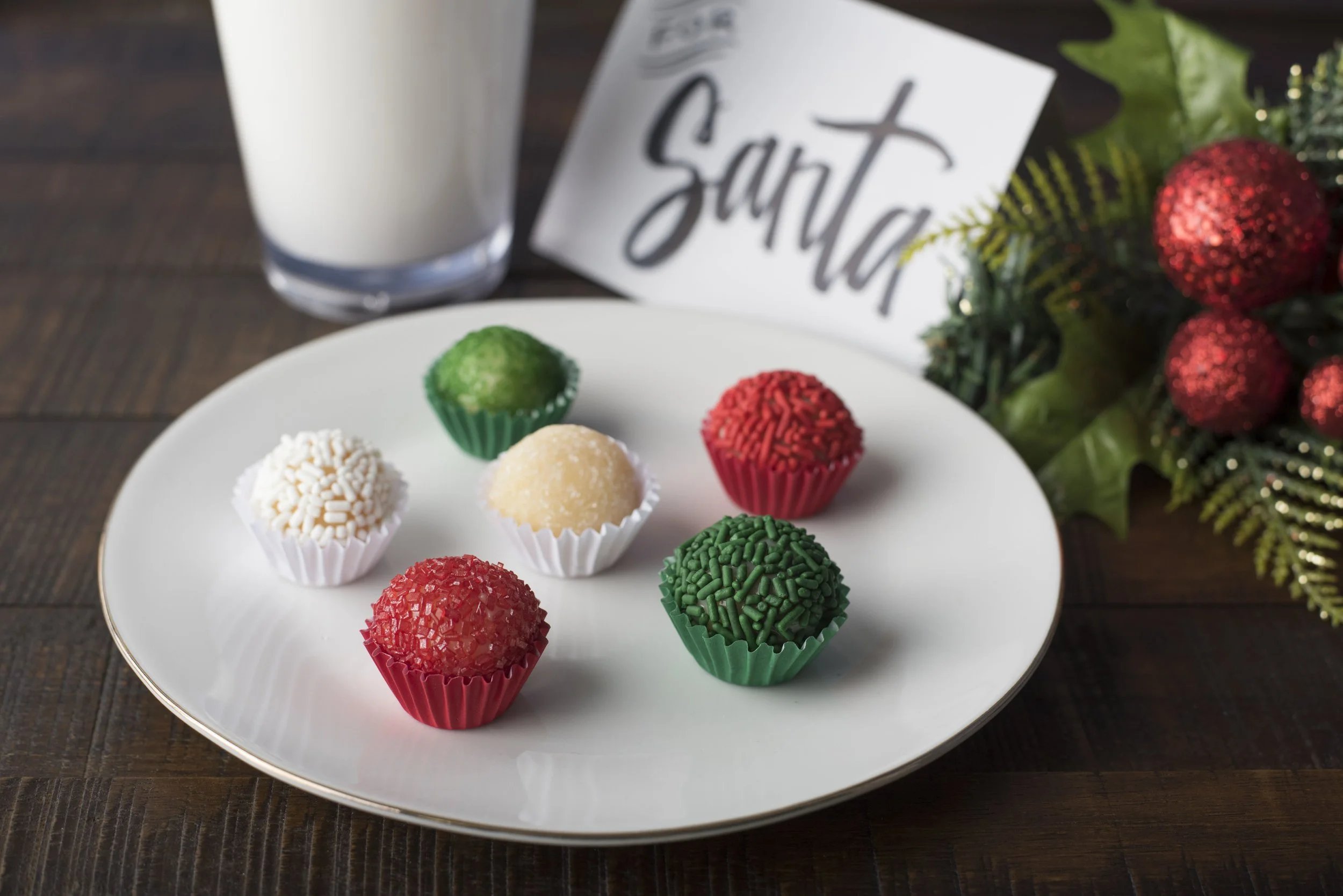 Assorted colorful truffles on a plate with a glass of milk, festive holiday decorations, and a card for Santa.