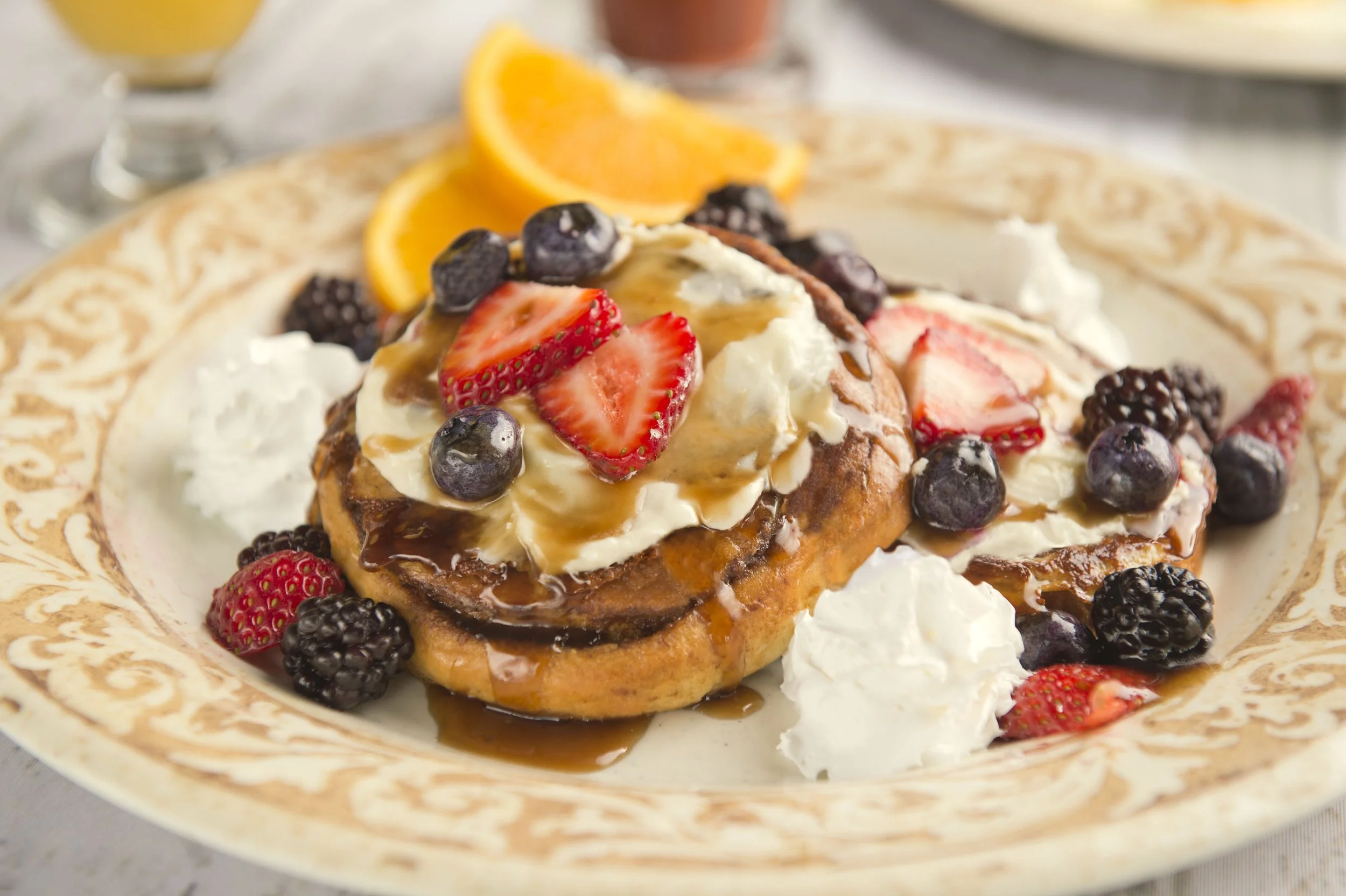 Plate of pancakes topped with whipped cream, strawberries, blueberries, blackberries, and syrup, with orange slices on the side.