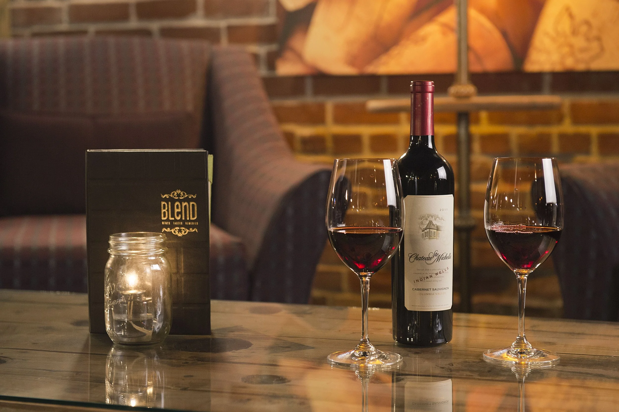 Two wine glasses filled with red wine, a bottle of red wine labeled Chateau Ste. Michelle, a lit candle in a glass jar, and a menu titled "Blend" on a wooden table with a cozy, dimly lit lounge setting in the background.