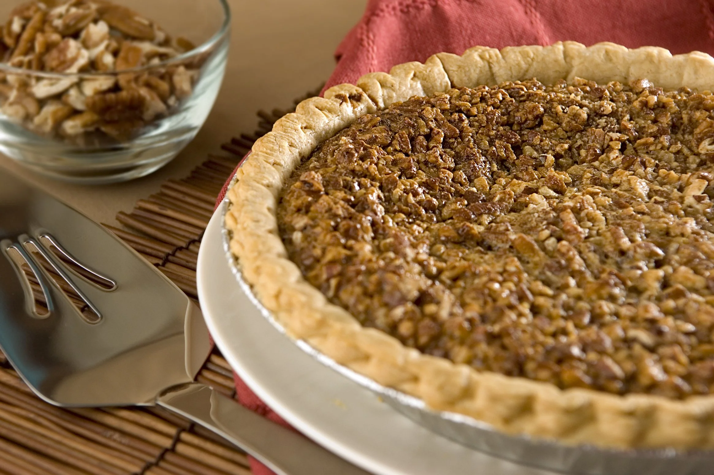 Pecan pie on a plate next to a bowl of pecans, with a pie server on the side.