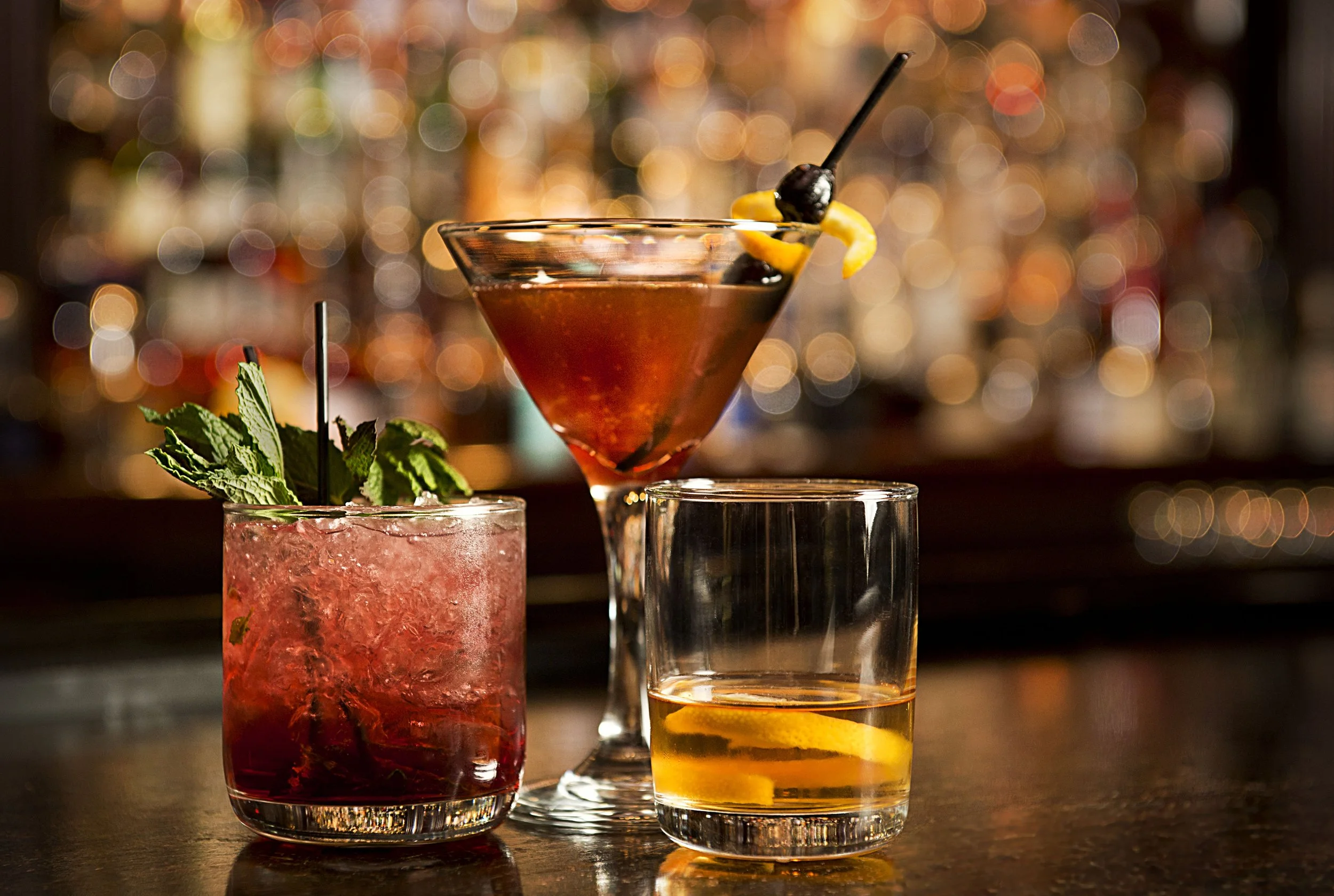 Three cocktail drinks on a bar counter with a blurred background; one with mint leaves, one in a martini glass with orange peel and a cherry, and one with a lemon wedge.