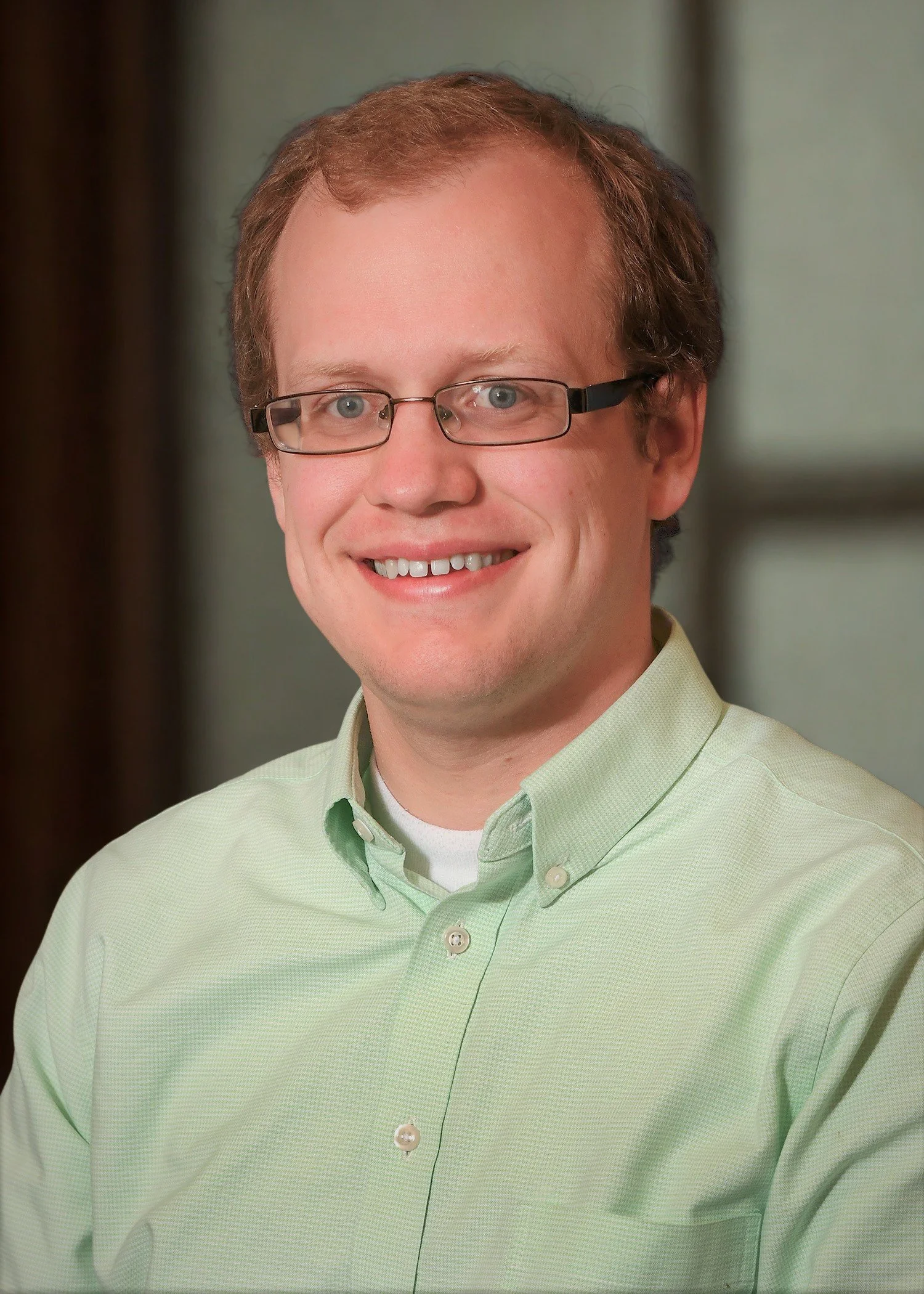 Headshot of a smiling man with short, light brown hair and glasses, wearing a light green button-up shirt, against a mottled grey background.