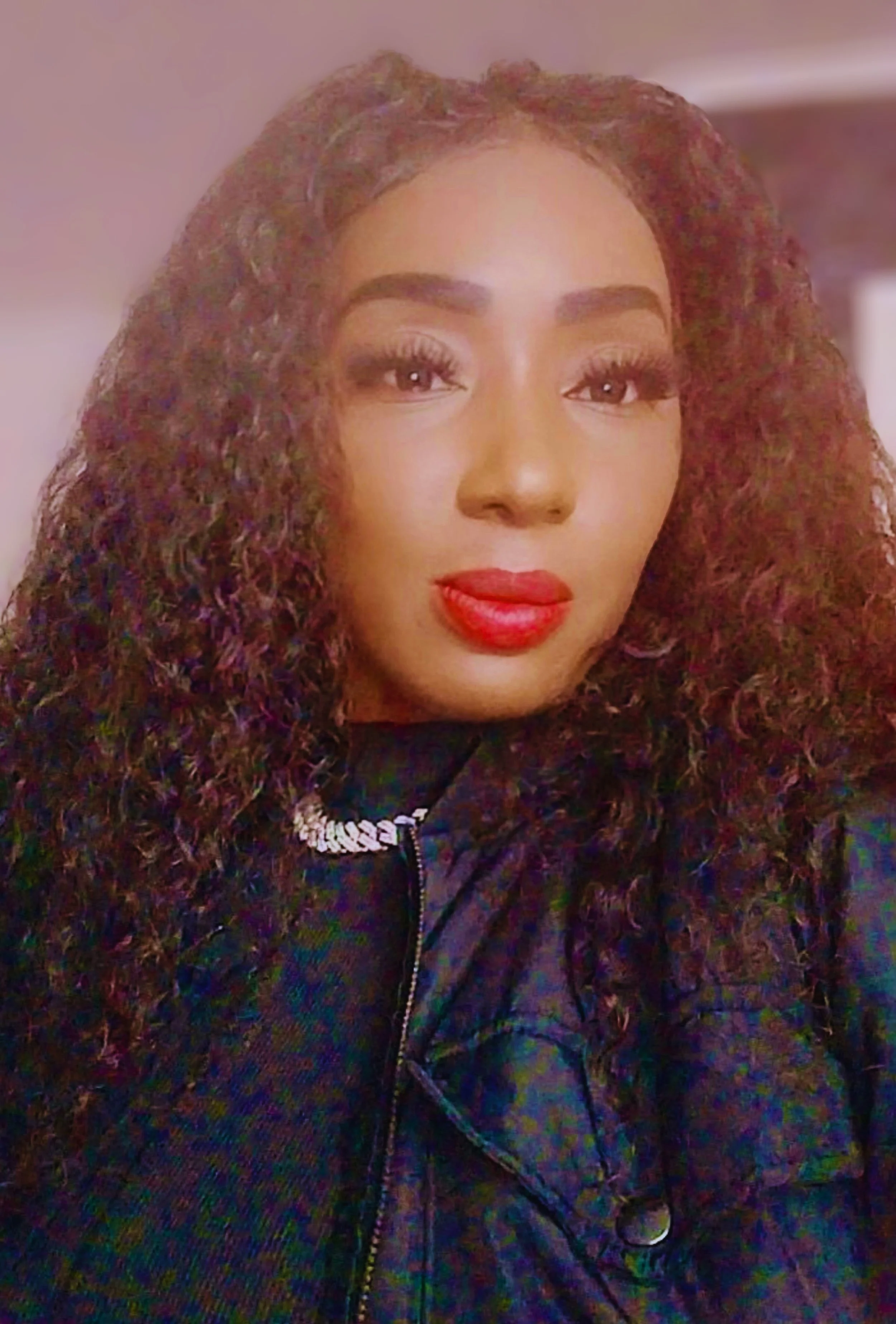 A woman with curly dark hair, bold red lipstick, and a silver chain necklace, wearing a black jacket, looking directly at the camera.