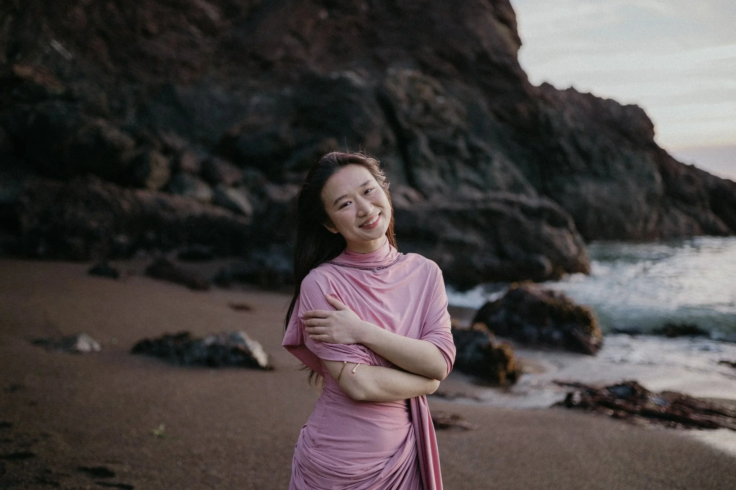 A young woman in a pink dress standing on a beach with rocky cliffs in the background, smiling at the camera during sunset.