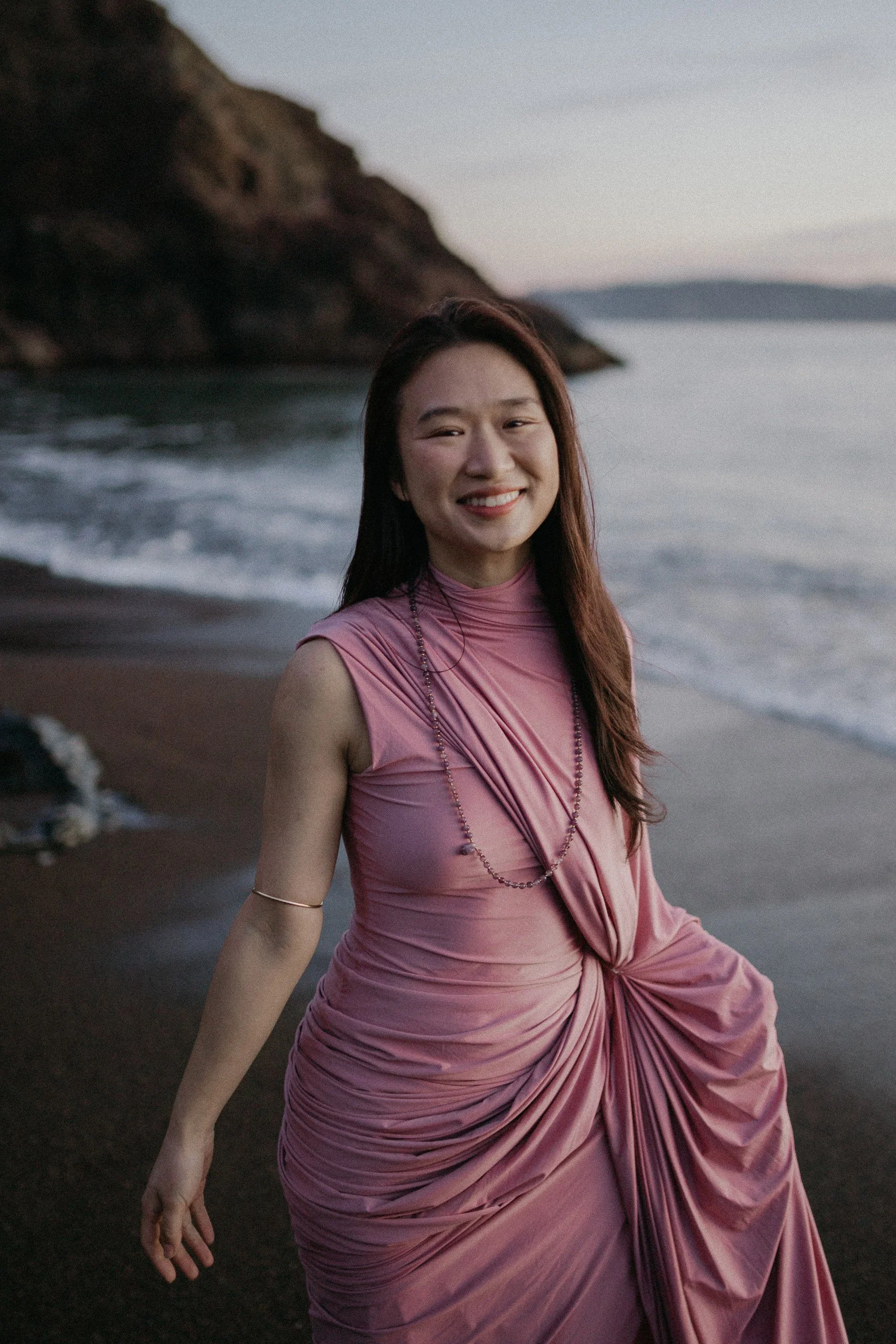 A woman in a pink dress smiling on a beach at dusk.