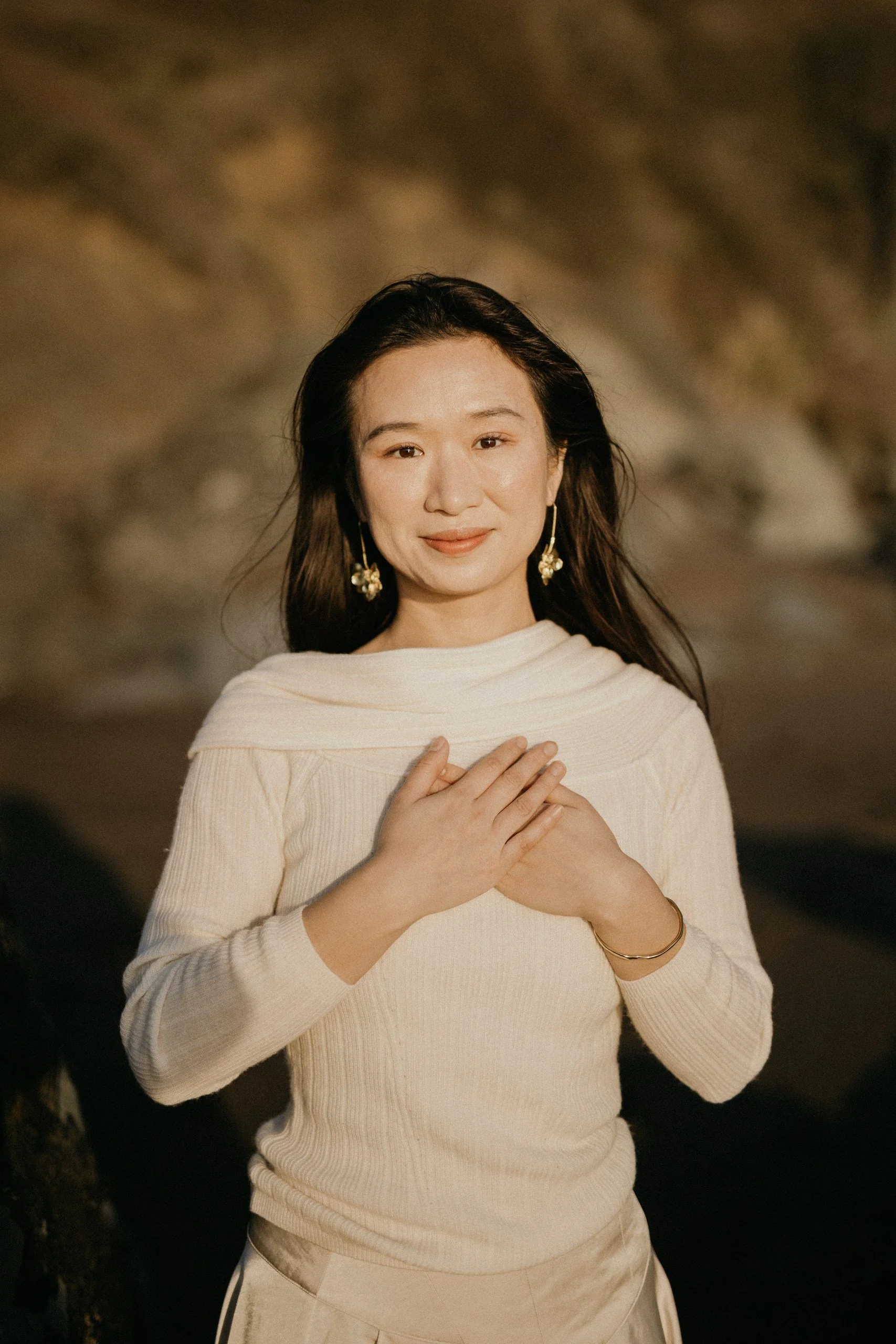 A woman with long dark hair, wearing earrings and a light-colored sweater, stands outdoors with her hands crossed over her chest, smiling softly at the camera during sunset.