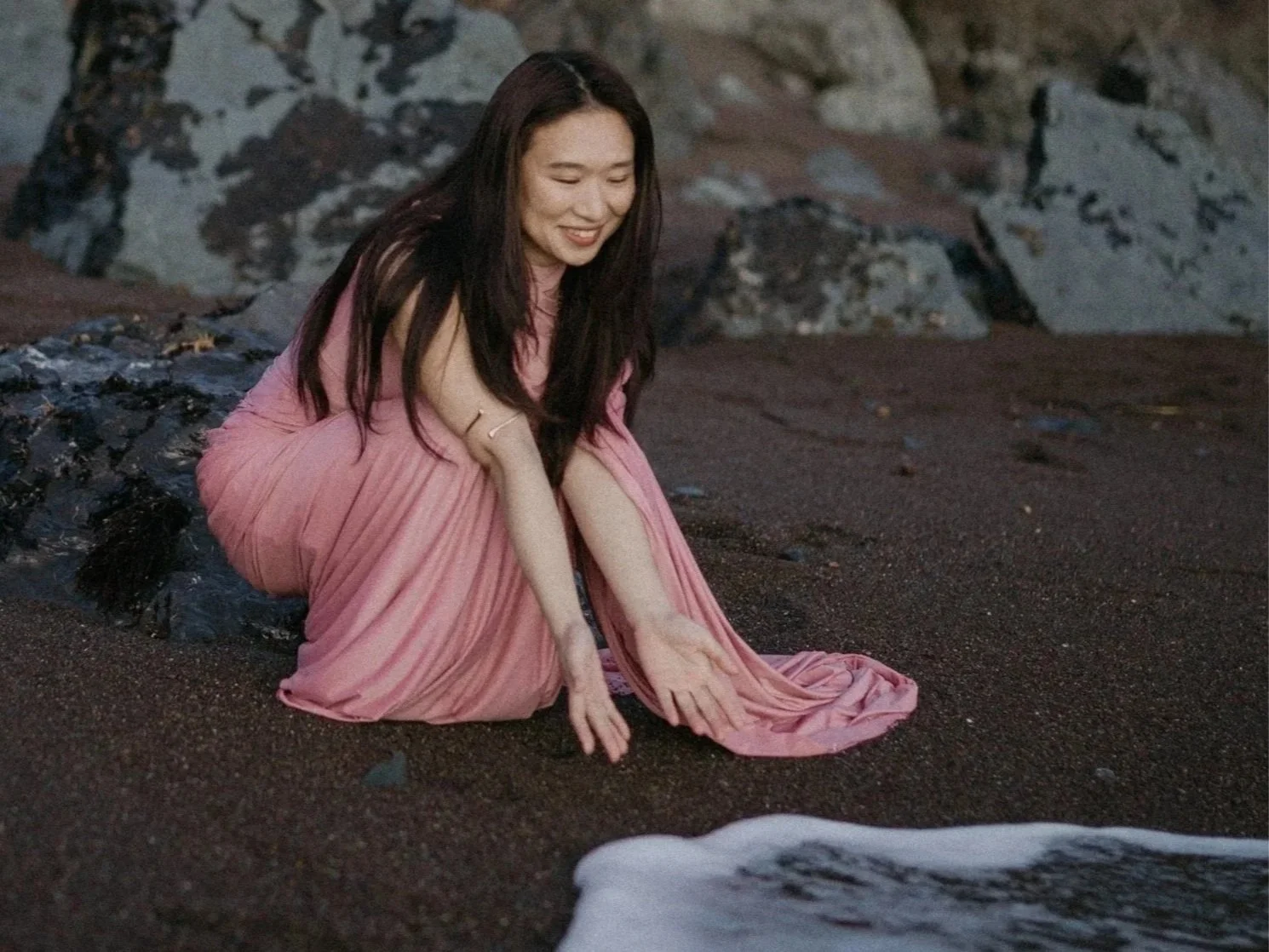A woman in a pink dress is sitting on the sandy beach near rocks, smiling and reaching out toward the water.