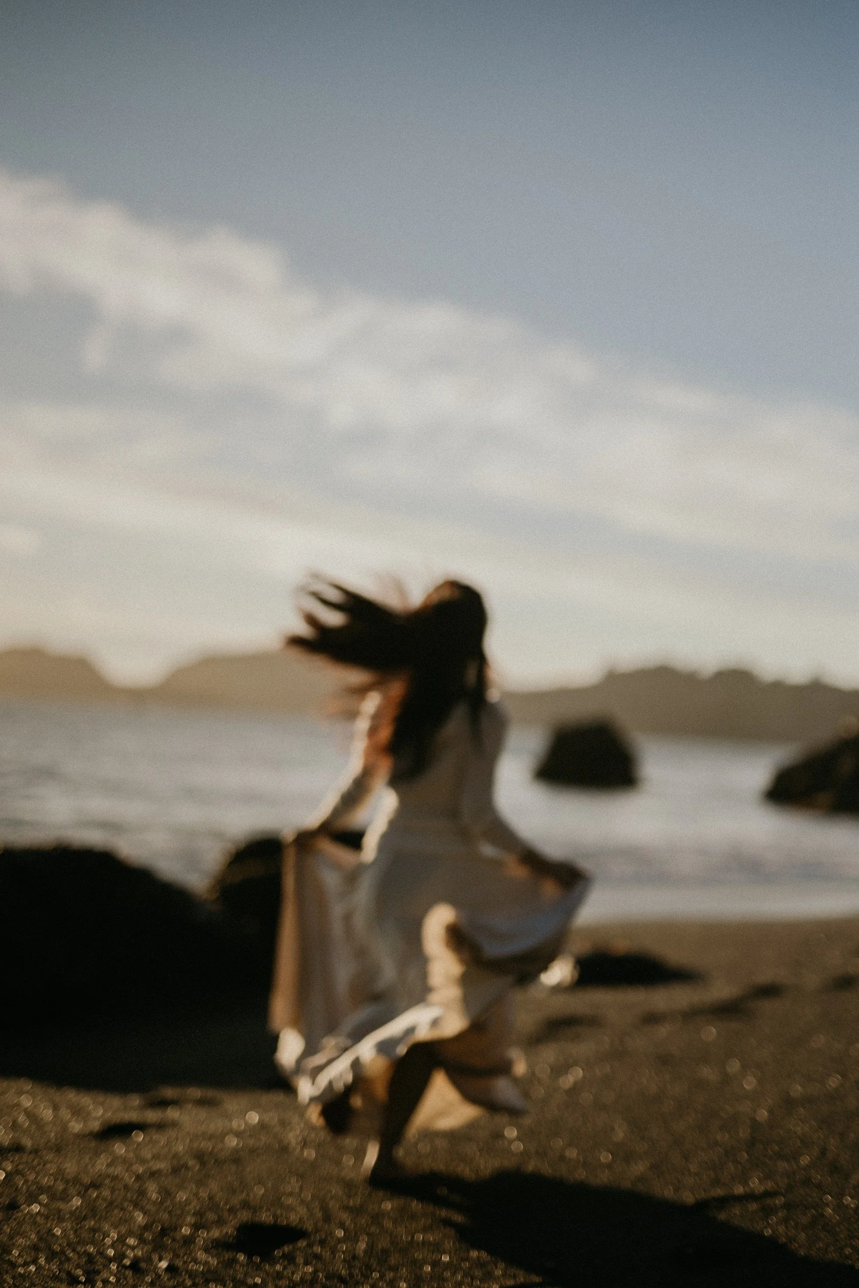 Woman in white dress sitting on a rock on the beach during sunset, with her hair flying in the wind and mountains in the background.