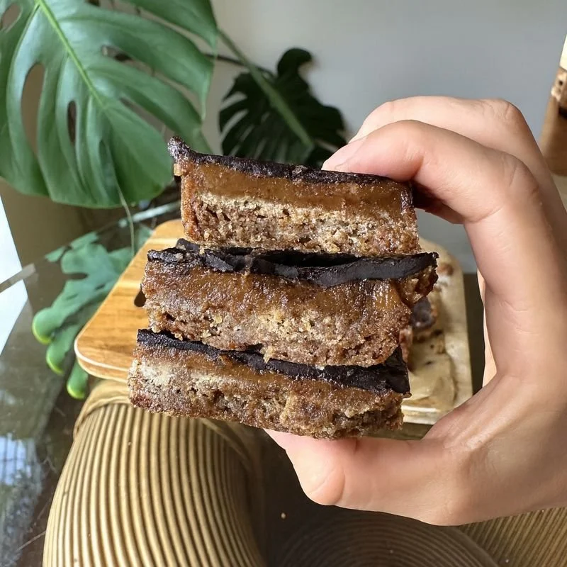 Three gingerbread caramel slices stacked on top of one another held in a person's hand.  The slices have a golden gingerbread base, a fudgey caramel centre and a dark chocolate top layer.