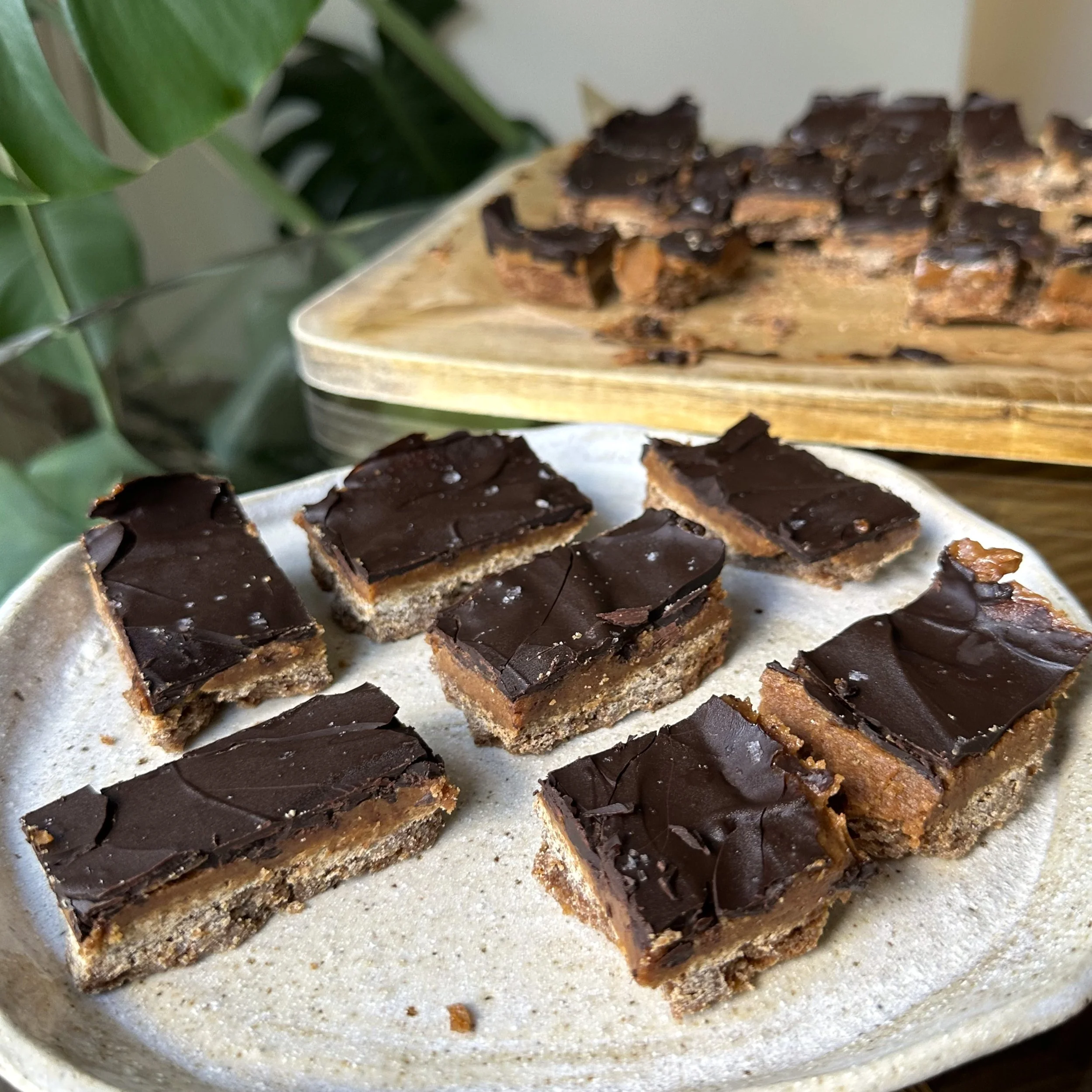 A ceramic plate is filled with gingerbread caramel slice. In the background is a wooden chopping board also filled with gingerbread caramel slice. The slices have a golden gingerbread base, a fudgy caramel centre, and a dark chocolate top layer.