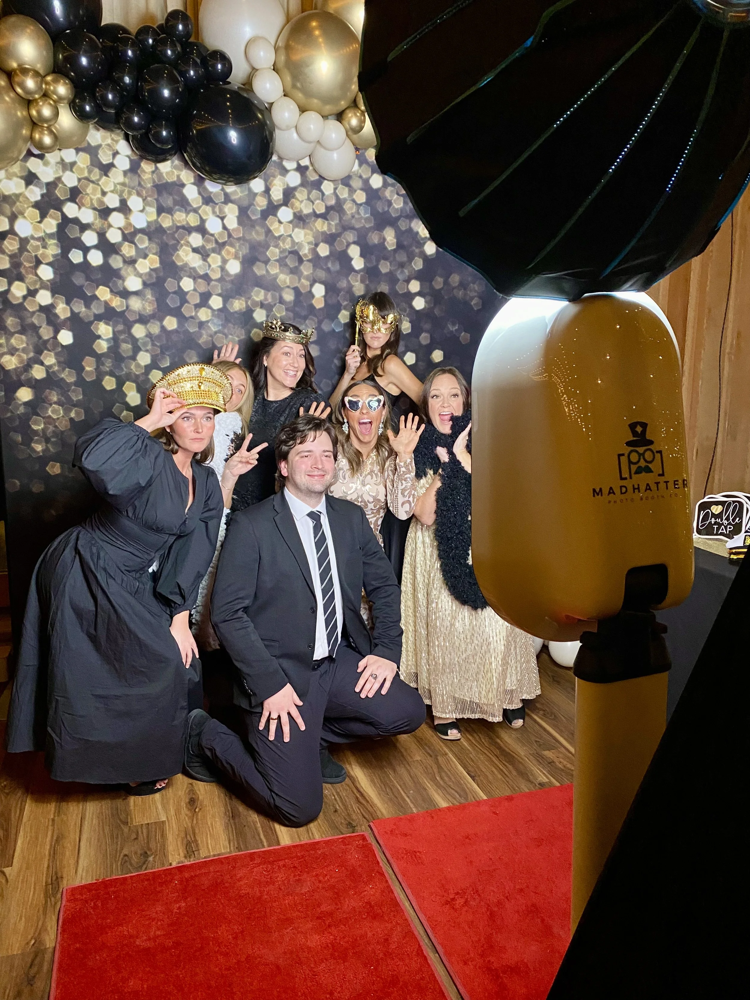Group of four people in formal wear posing and making playful expressions in front of a photo booth with a gray backdrop.