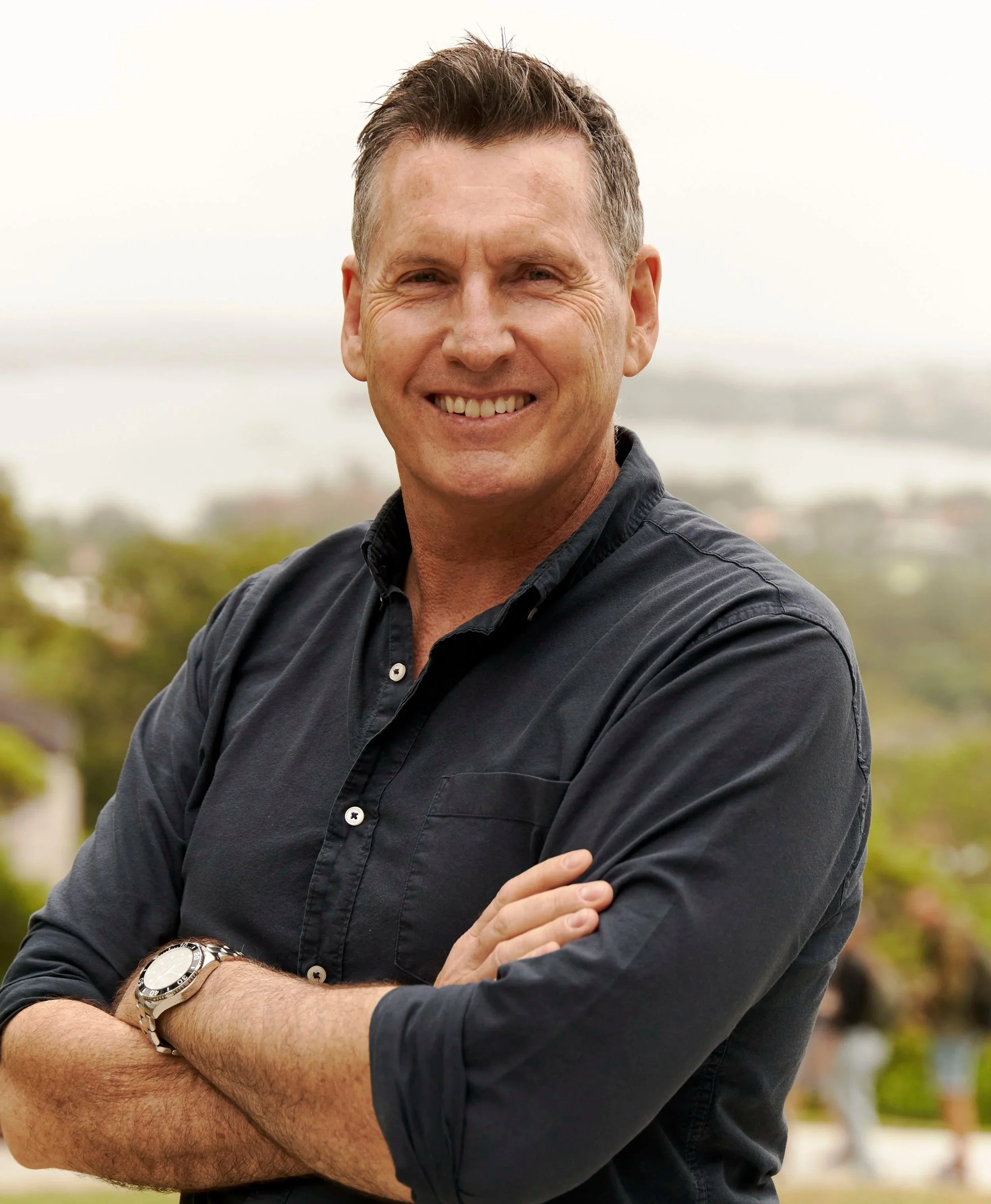 A smiling man with short brown hair, wearing a black button-up shirt with his arms crossed, standing outdoors with a blurred background of trees and a cloudy sky.
