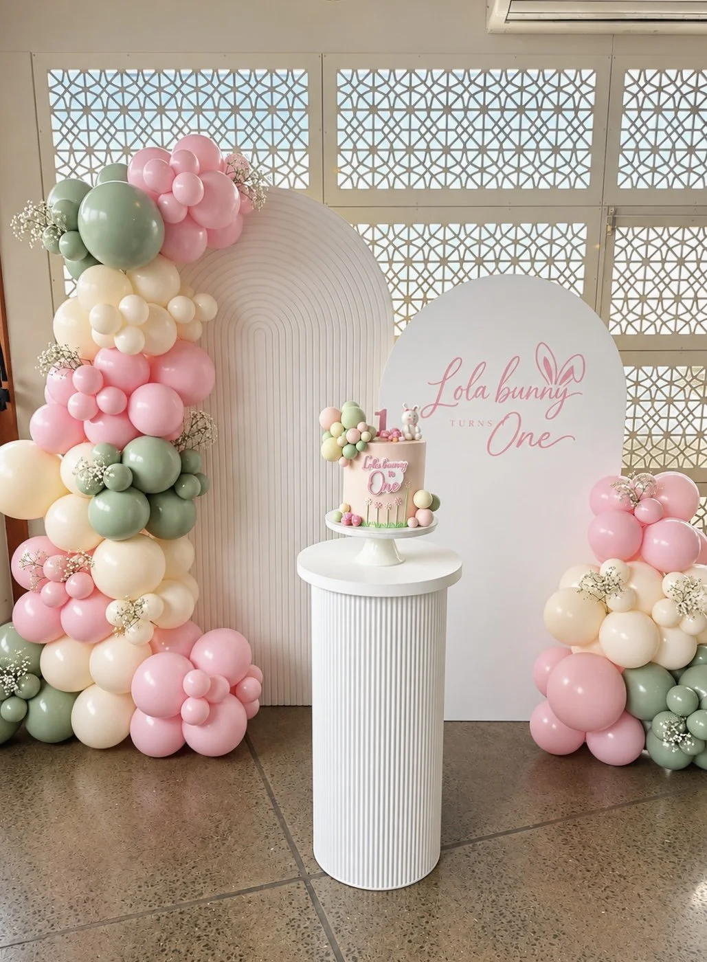 Decorative backdrop with pastel-colored balloons on either side of a white and pink sign that reads 'ONE SILLY GOOSE,' with a small cake on a white pedestal in front, set on green artificial grass.
