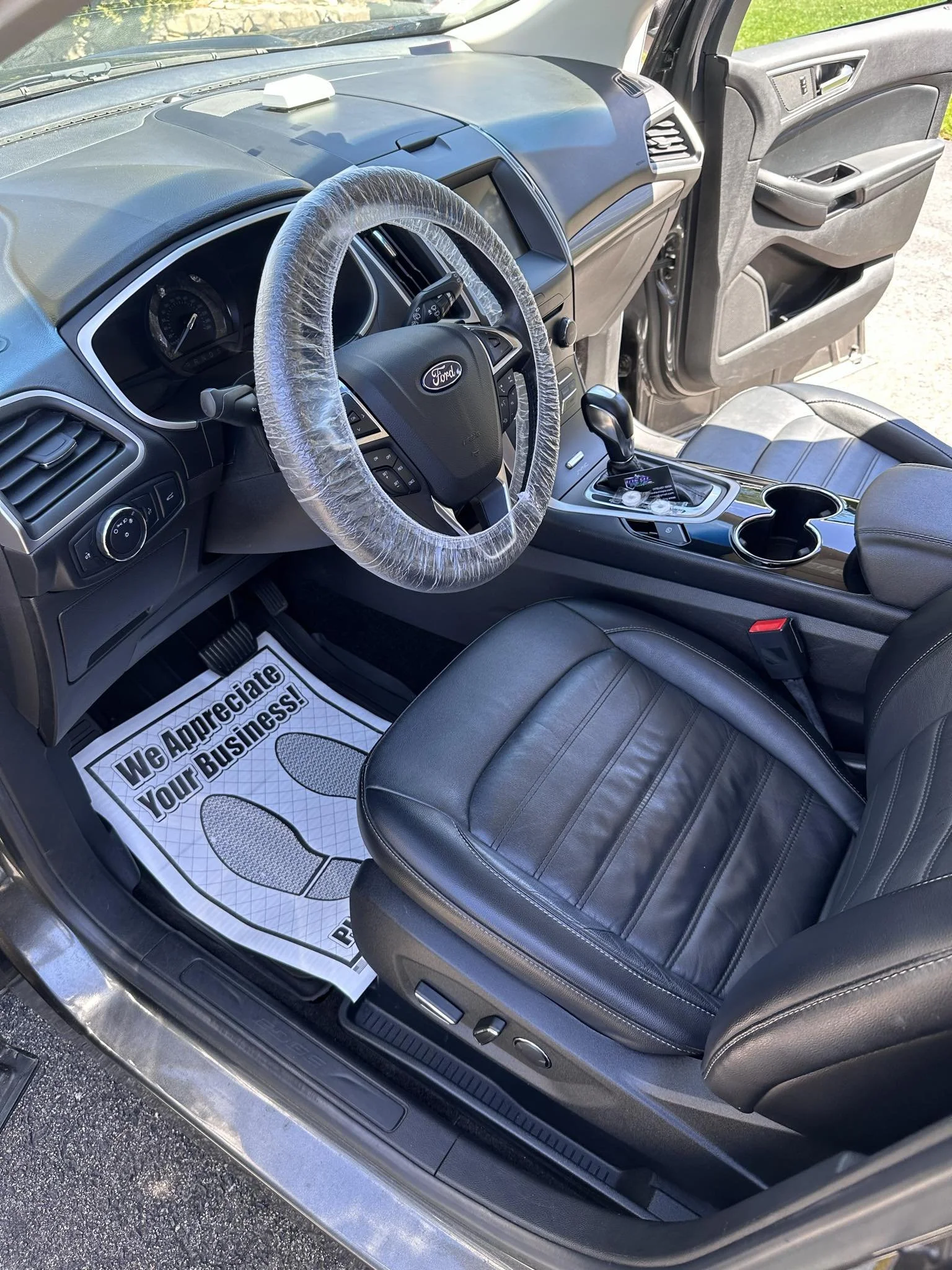 Interior of a Ford car with a covered steering wheel, leather seats, and a floor mat saying, "We Appreciate Your Business."