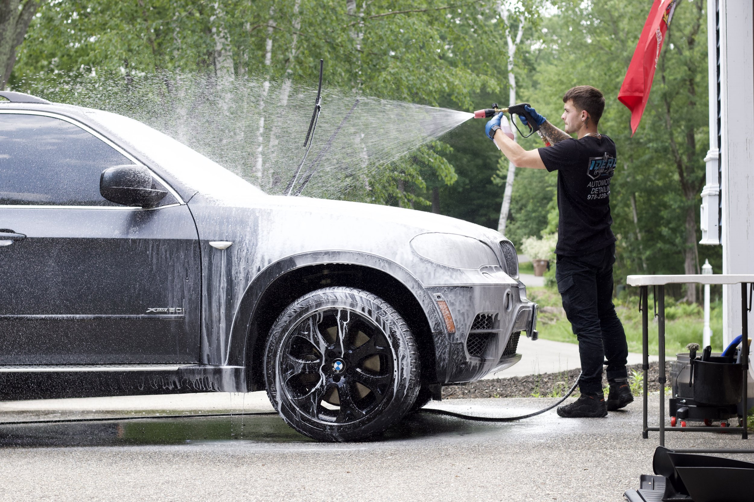 Man washing a black BMW car with a hose, with soap suds on the car, in an outdoor setting surrounded by green trees.