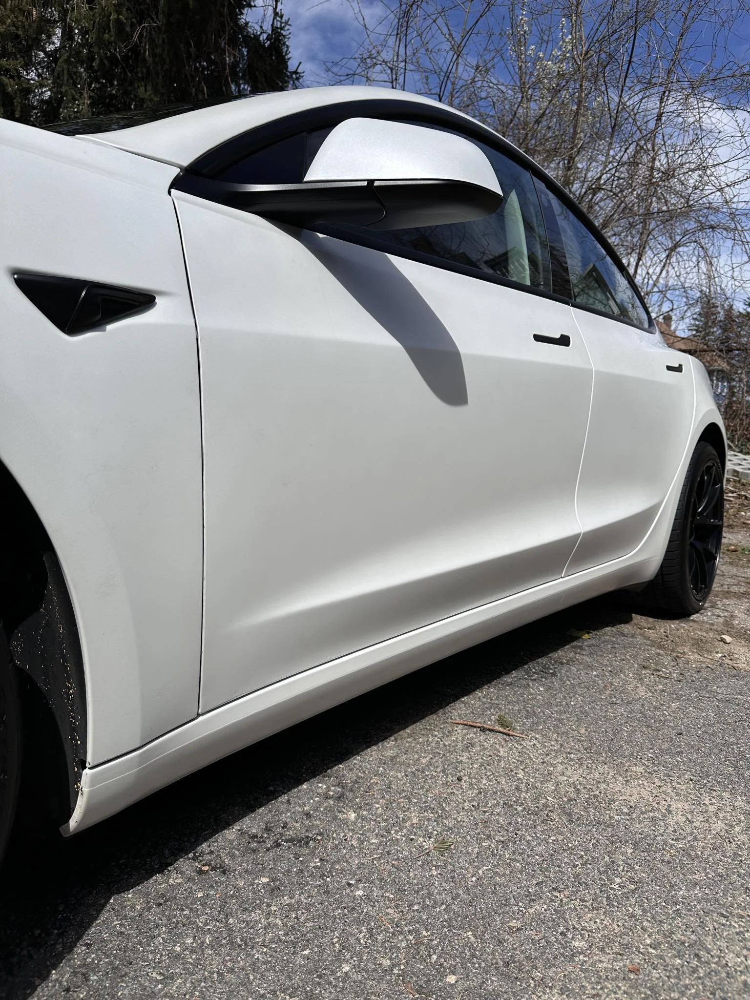 White car parked on a road, close-up of side view with black wheel, side mirror, and door handle, trees and sky in background.