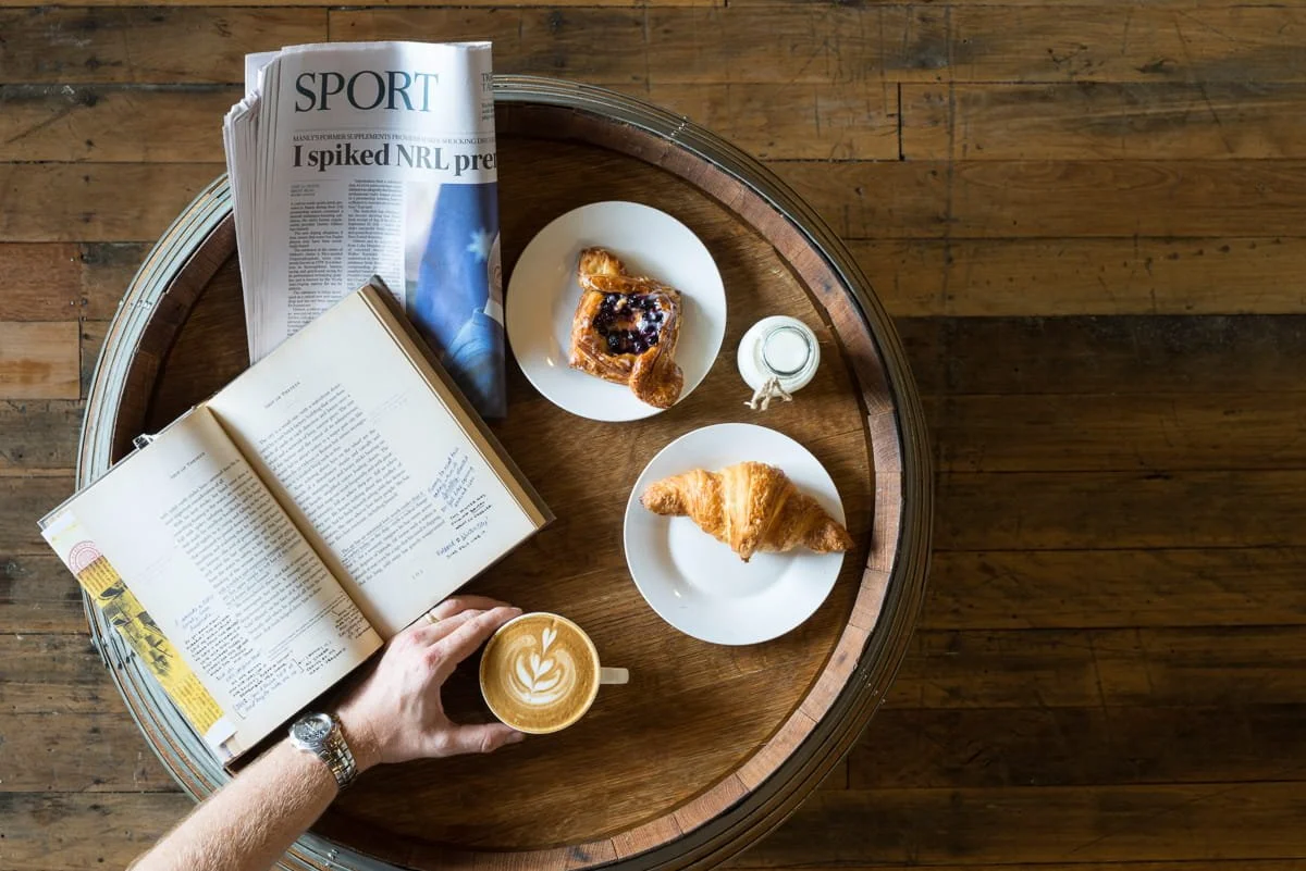 A commercial lifestyle image of a person's hand holding a coffee with a newspaper on the table. Taken by commercial photographer Tom MacDonald.