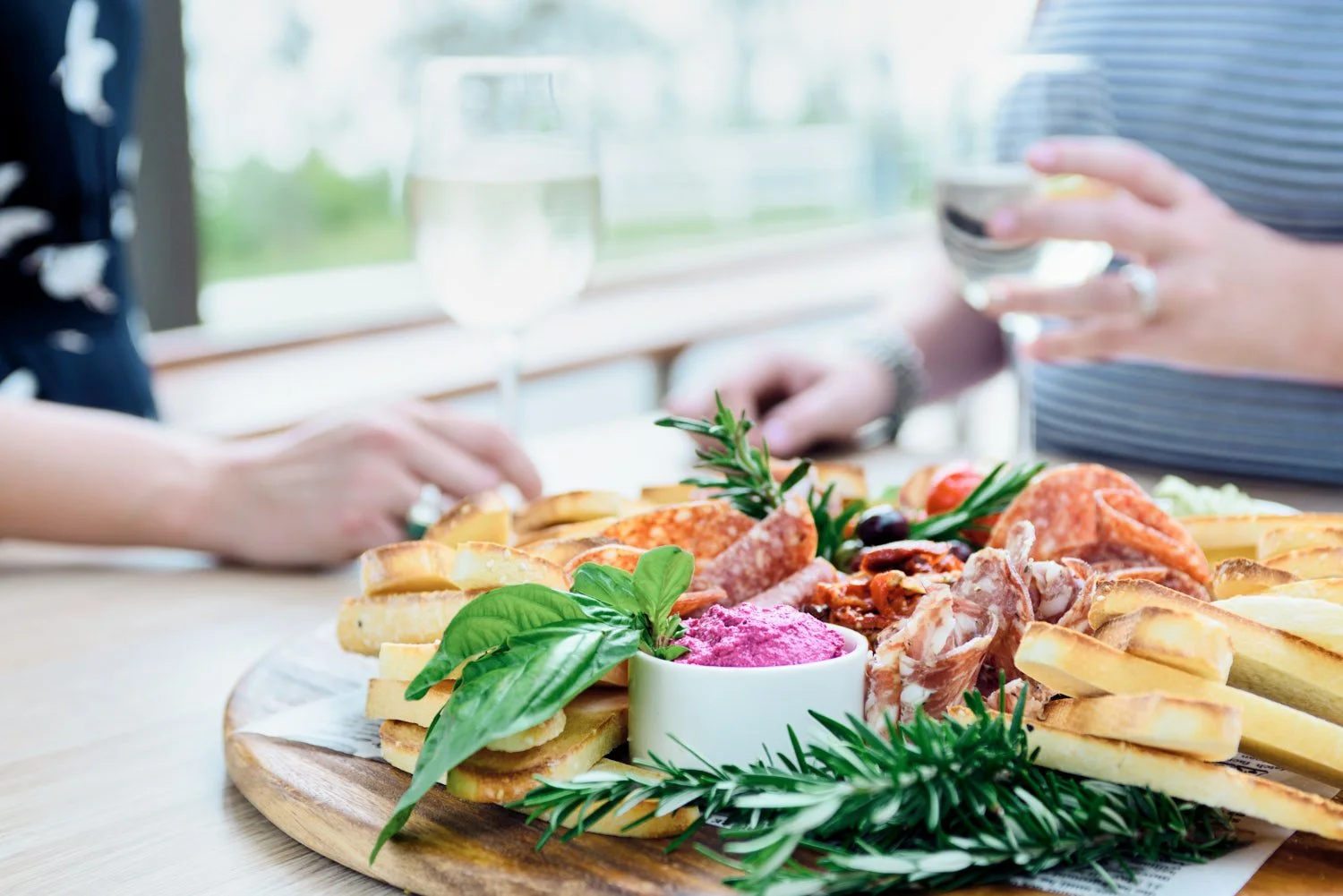 A restaurant photograph of acharcuterie board with bread, cheese, cured meats, and herbs, with two people enjoying drinks, taken by Tom MacDonald of commercial photography company Tom Mac Photography.