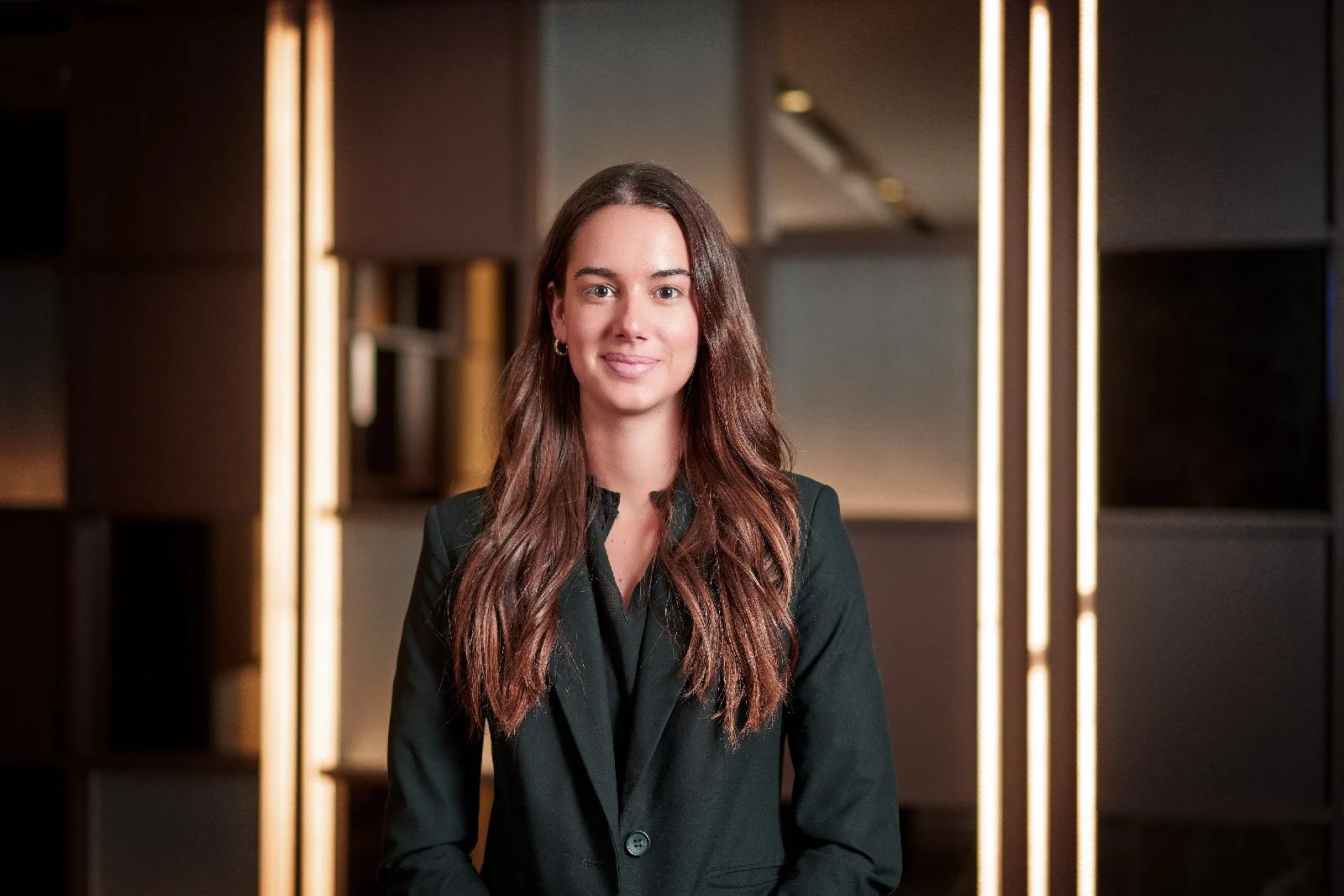 A n environmental headshot of young woman with long, wavy brown hair wearing a black blazer, standing in front of a modern, dimly-lit office space with vertical light strips and bookshelves. Taken in Brisbane by Tom MacDonald of TomMac photography