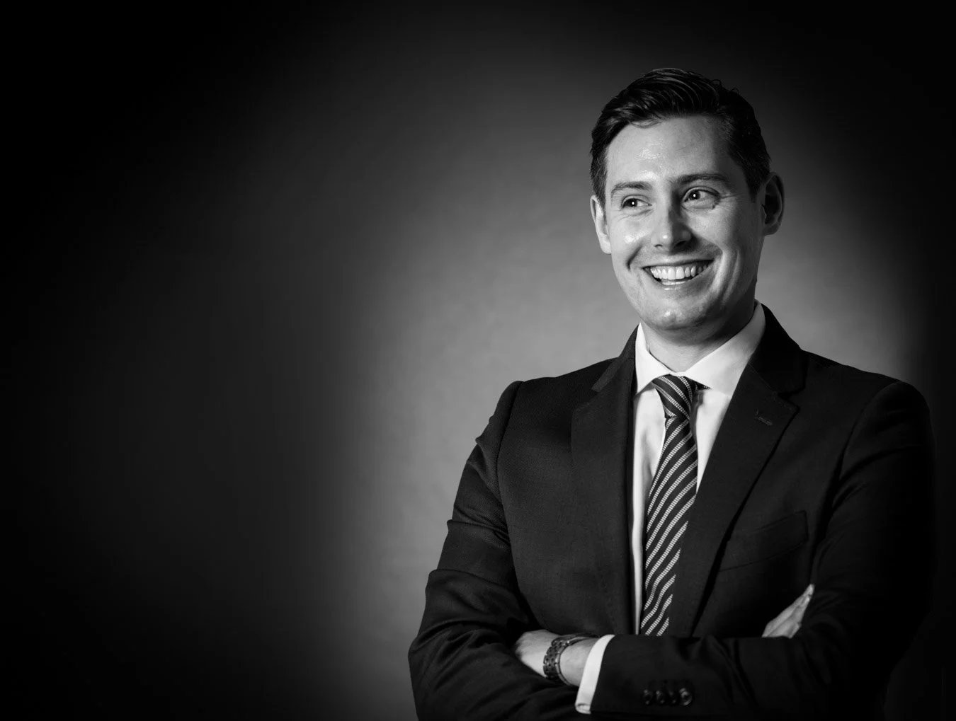 Black and white studio portrait of a smiling young man in a suit with arms crossed, facing slightly right. Taken in Brisbane by Tom MacDonald