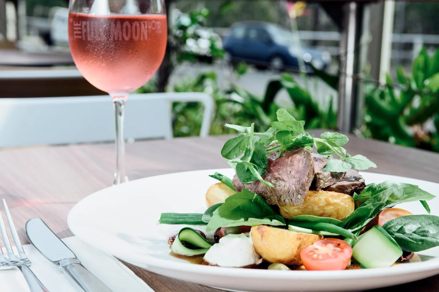 A  commercial photograph of a plate of food with grilled steak, tomatoes, cucumbers, and greens, next to a glass of rosé wine, on a wooden table outside. Taken by Brisbane commercial food photographer Tom Mac photography.