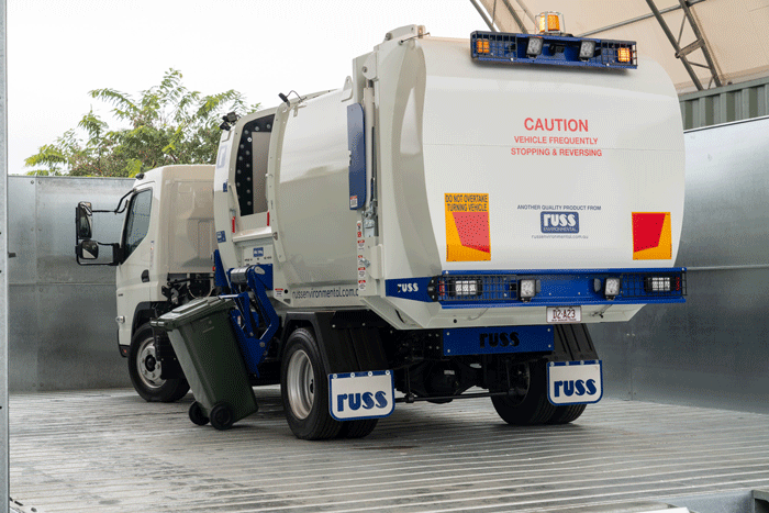 A  gif created by Tom MacDonald of astreet sweeper truck parked on a metallic surface in Brisbane, equipped with a large white tank and blue accents, with warning signs and a green trash bin attached.