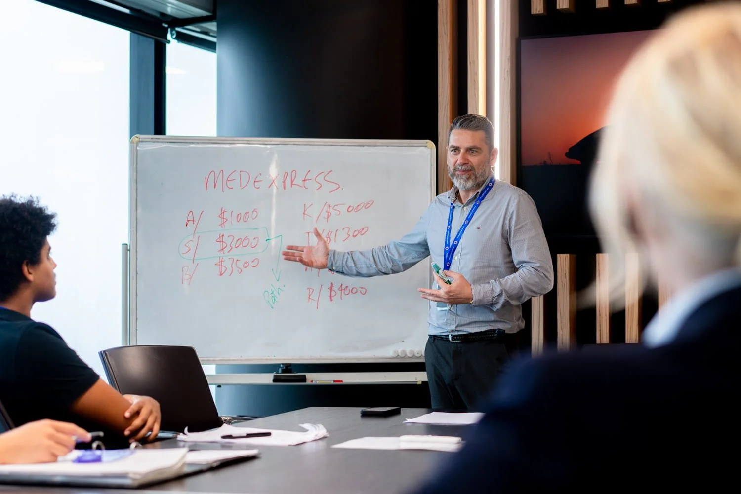 Man presenting in front of a whiteboard with diagrams and numbers, seated audience members in a meeting room with wood wall accents and a large monitor. Taken by commercial photographer Tom MacDonald in Brisbane.