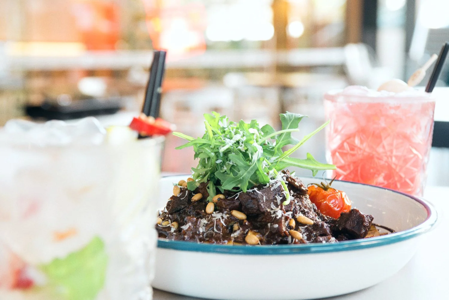 A commerciasl food imahe of a bowl of beef topped with greens and pine nuts, two drinks with straws in the background, in a bright casual restaurant or cafe setting, taken by Tom MacDonald of commercial photography company Tom Mac Photography.