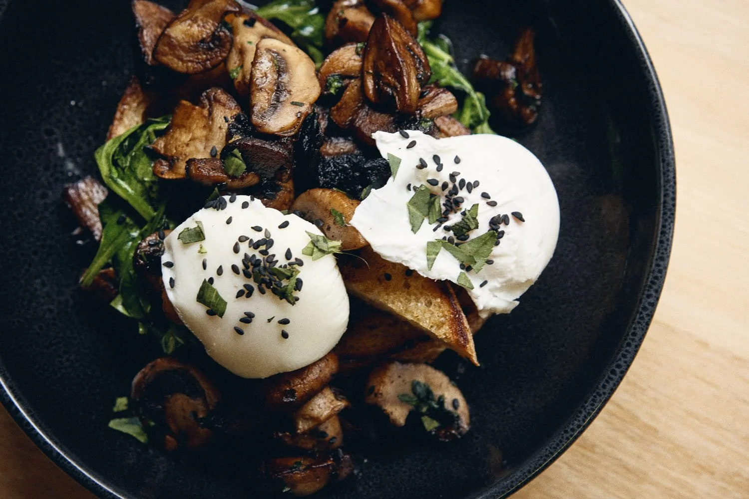 A  commercial food and restaurantt image of black bowl containing roasted mushrooms, two poached eggs topped with black sesame seeds, taken by Brisbane based photographer Tom MacDonald