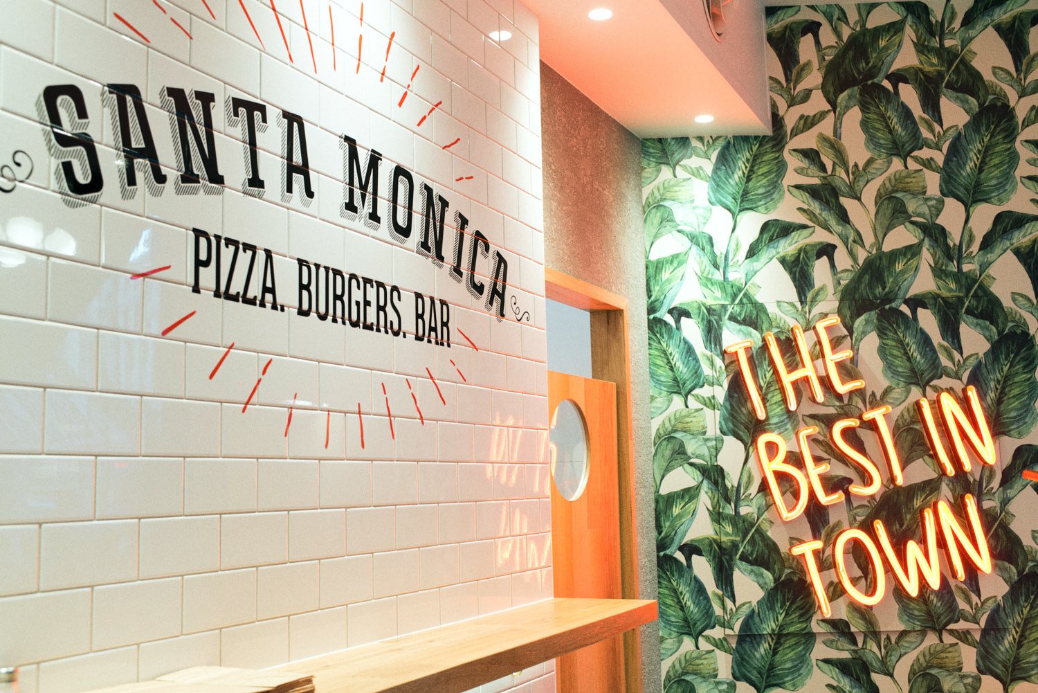 A commercial interior photograph of a restaurant with white subway tile wall displaying the name Santa Monica and the description Pizza, Burgers, Bar. Taken by Brisbane based commercial photographer Tom MacDonald.