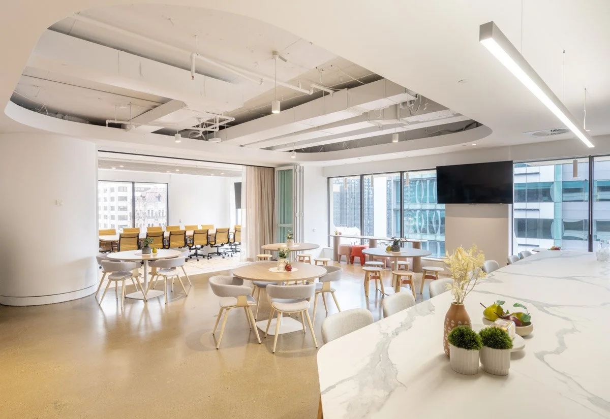 A commercial image of a Modern office break room with round tables, chairs, a large marble countertop, wall-mounted TV, large windows, and cityscape view. taken by Tom MacDonald