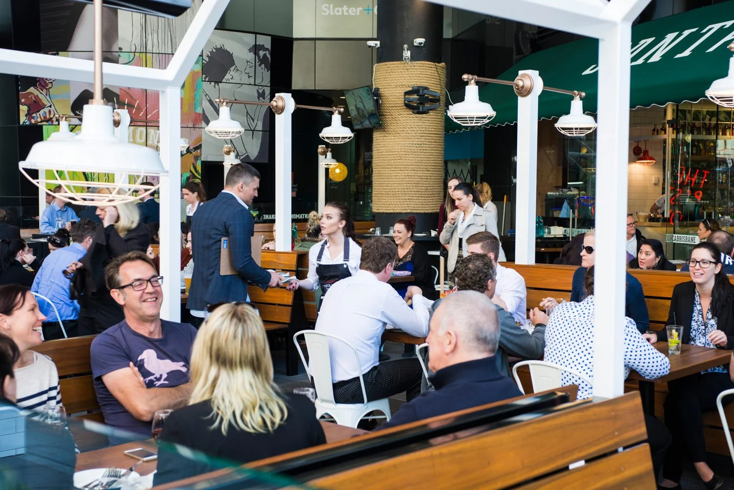 A commercial image taken by Tom MacDoanld of people dining at an outdoor Brisbane restaurant, some are smiling and talking, with a server handing a guest a dish, and a woman taking a photo, under green and white umbrellas, behind a glass barrier.