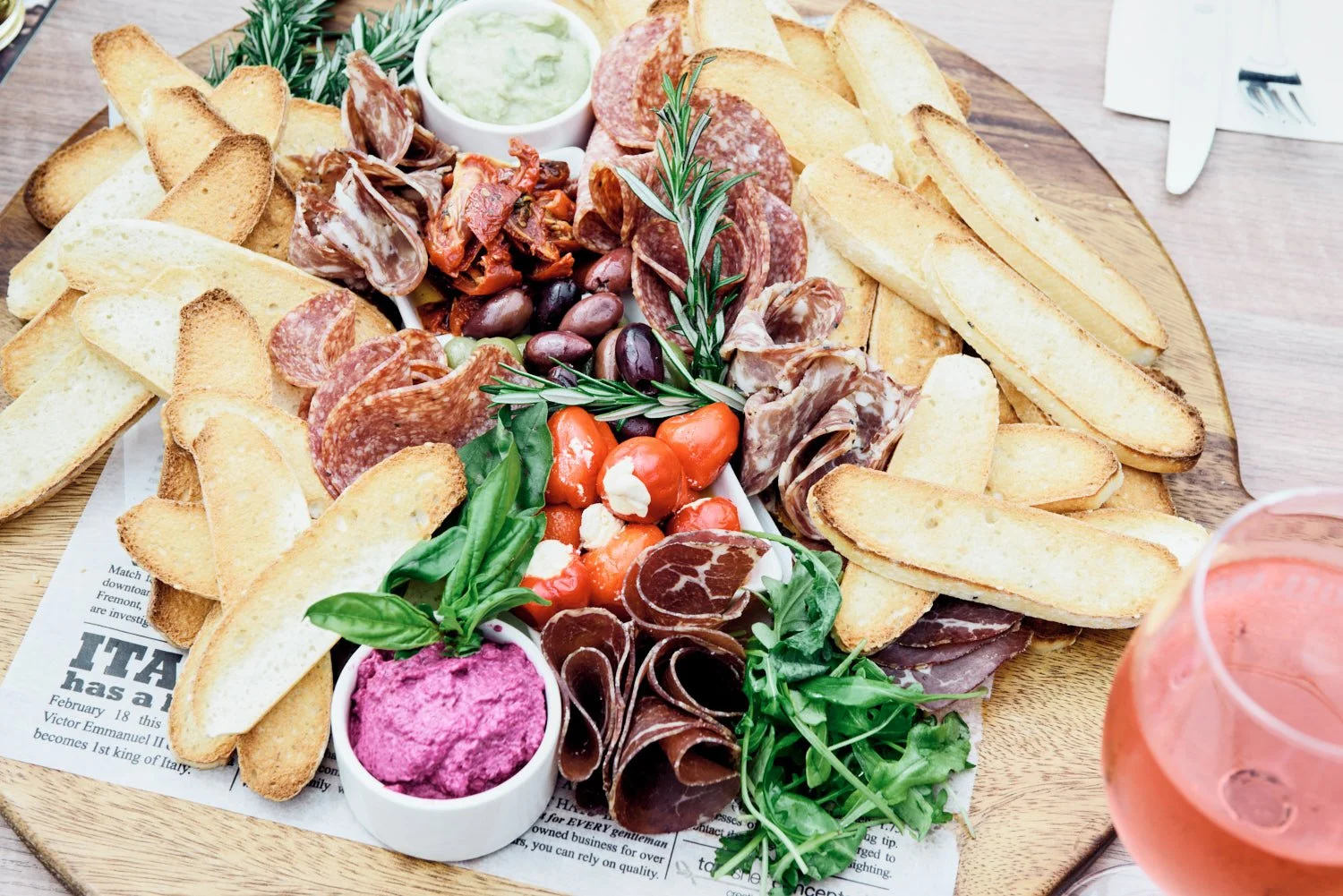 A photo of a wooden platter with an assortment of sliced bread, cured meats and olives, Taken by Commercial food photographer Tom MacDonald of Tom Mac photography.
