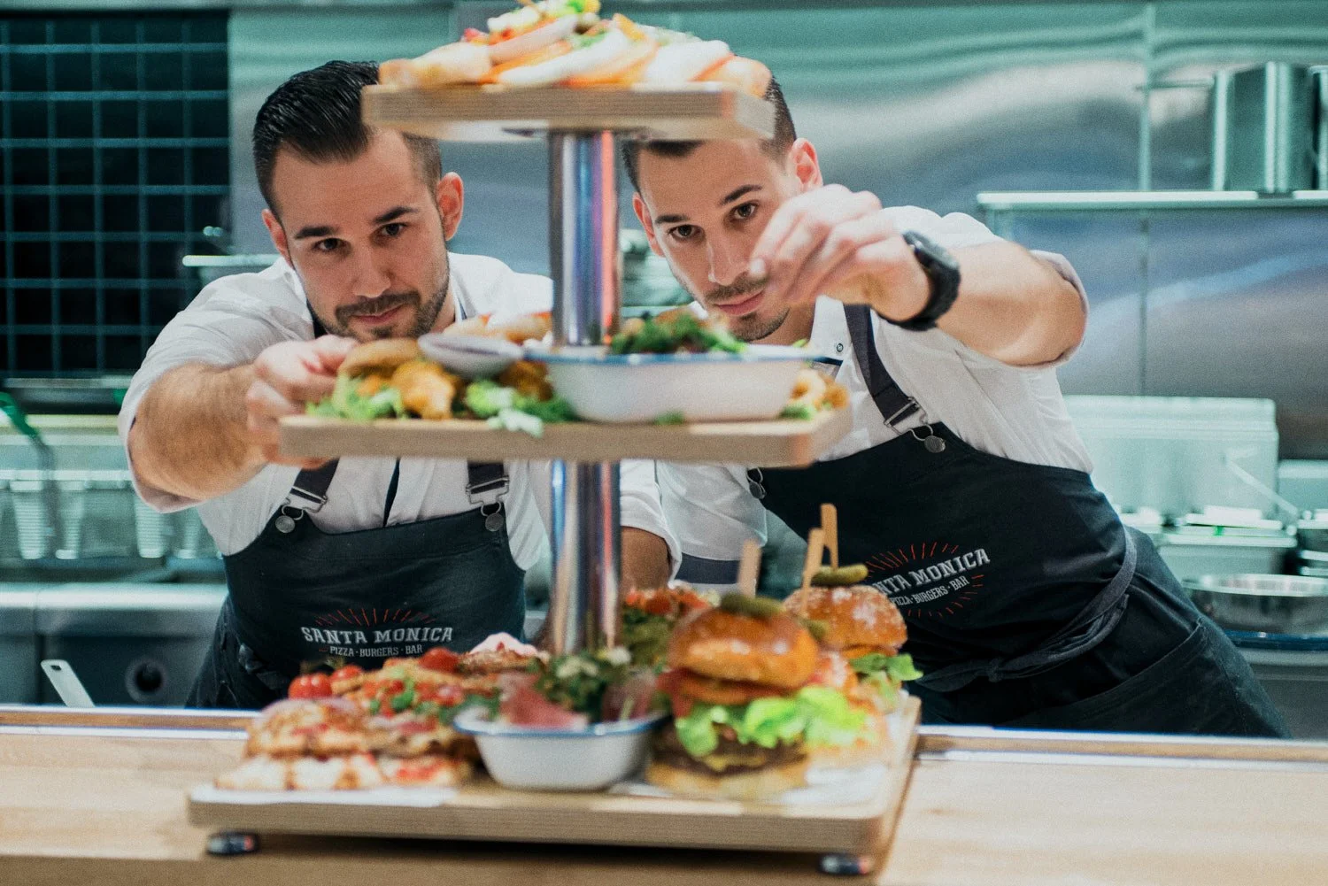 A commercial photo of two chefs in black aprons preparing a multi-level tray of food, including burgers and appetizers, in a professional kitchen. Taken by food photographer Tom MacDonald of Tom Mac photography.