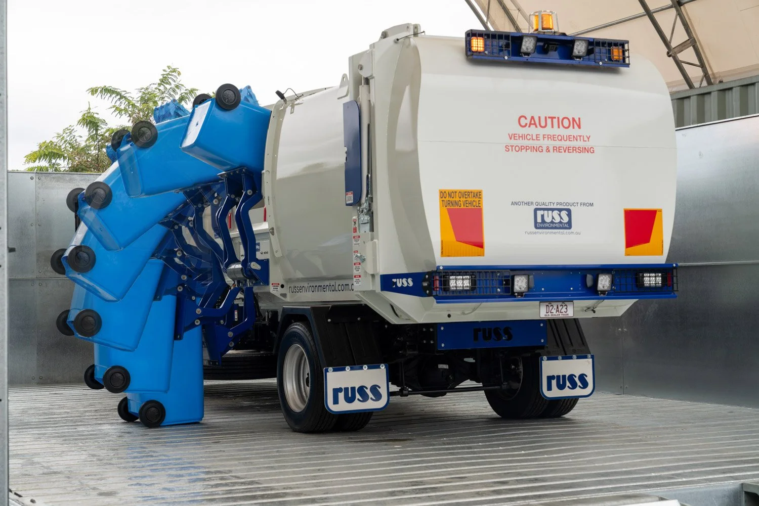  A photograph taken by Brisbane based Tom MacDonald of Tom Mac photography,  of a street cleaning vehicle with blue water tanks on the side, white body, and blue accents, parked inside a metallic enclosure. 