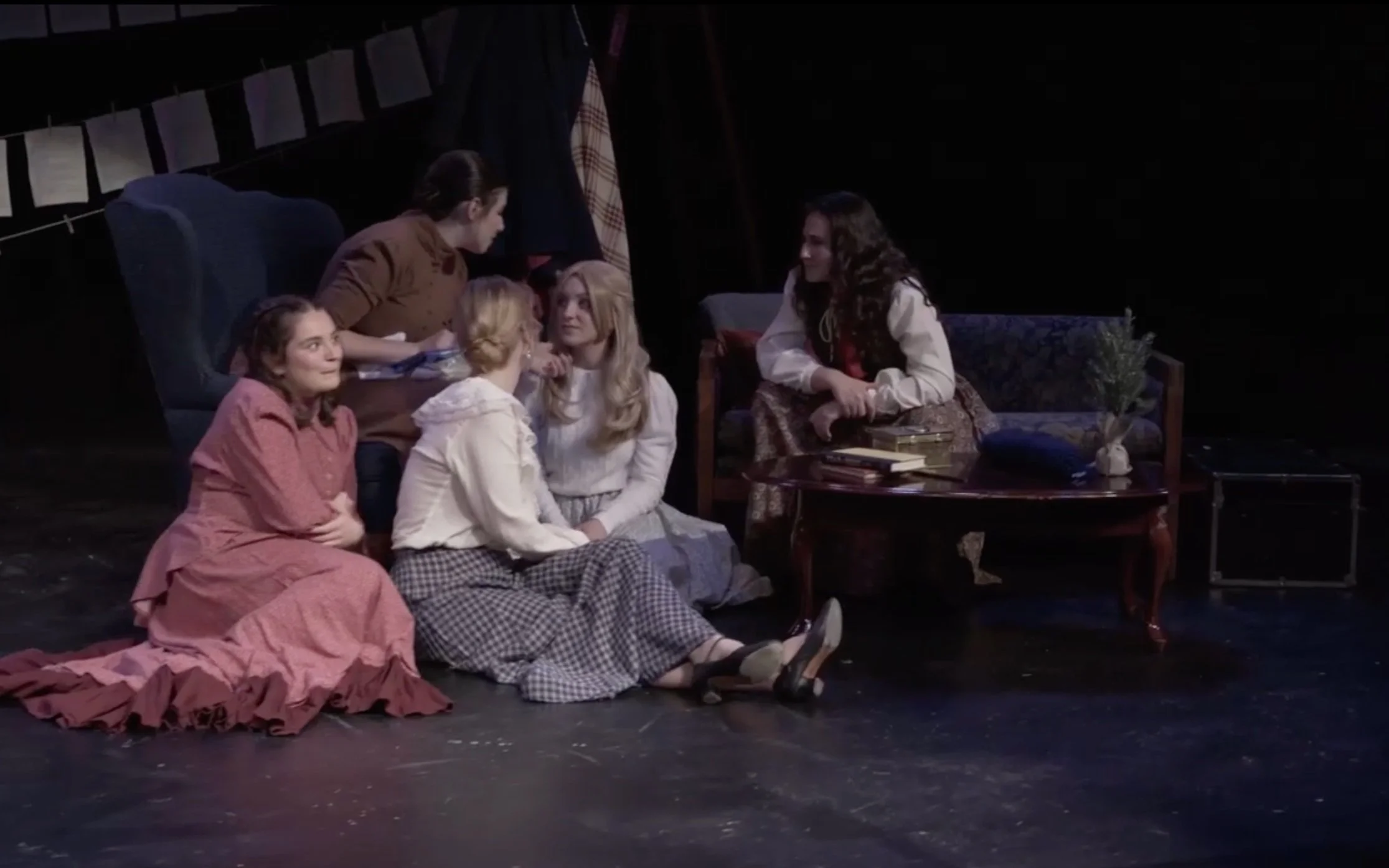 The girls watch Marmee open her presents. A variety of set dressing props can be seen on the coffee table, and in the shadows on the right side sits the chest of costumes.