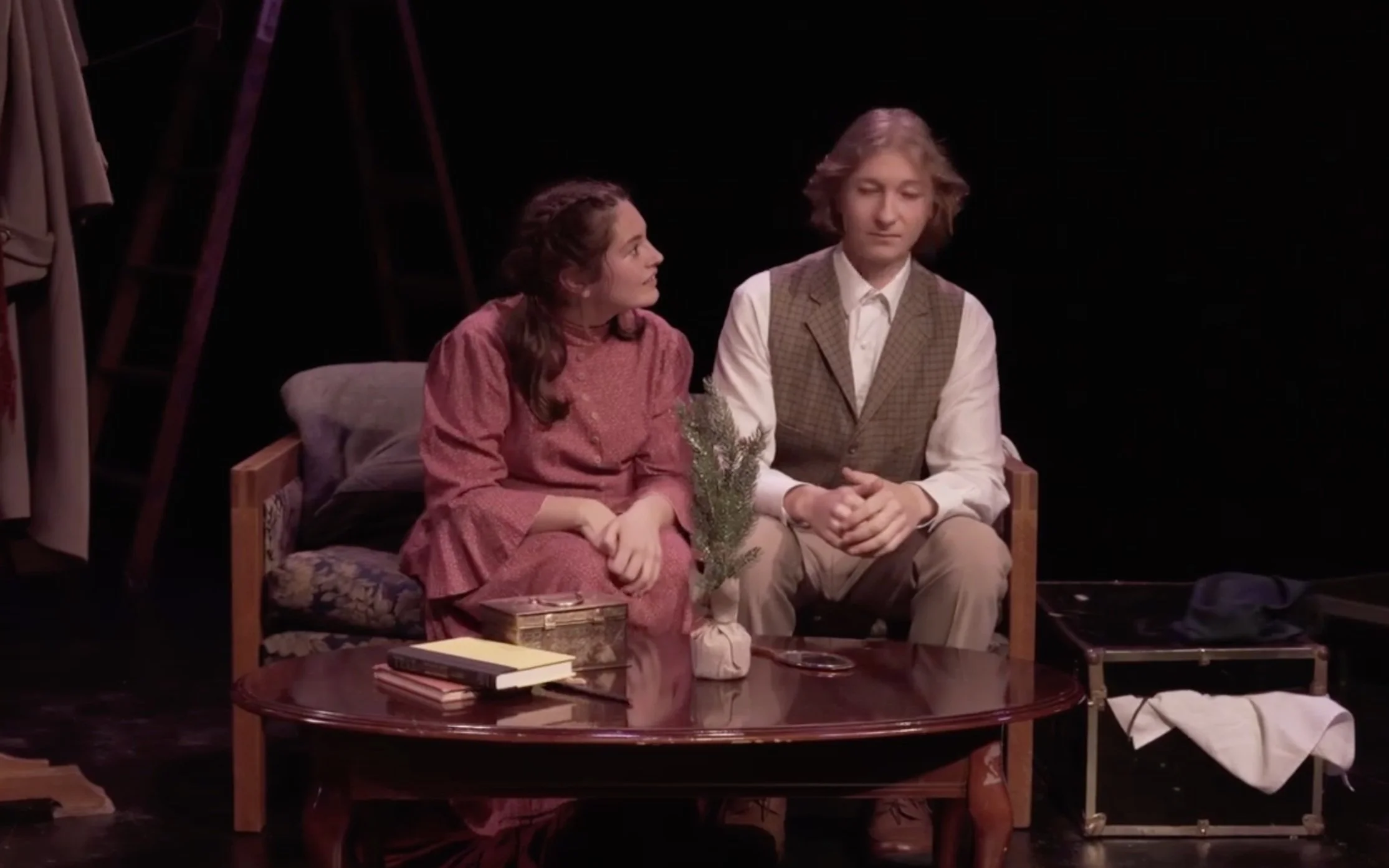 Laurie sits with Amy. Hand and dressing props can be seen on the table, and the chest of costumes is in the bottom right corner.