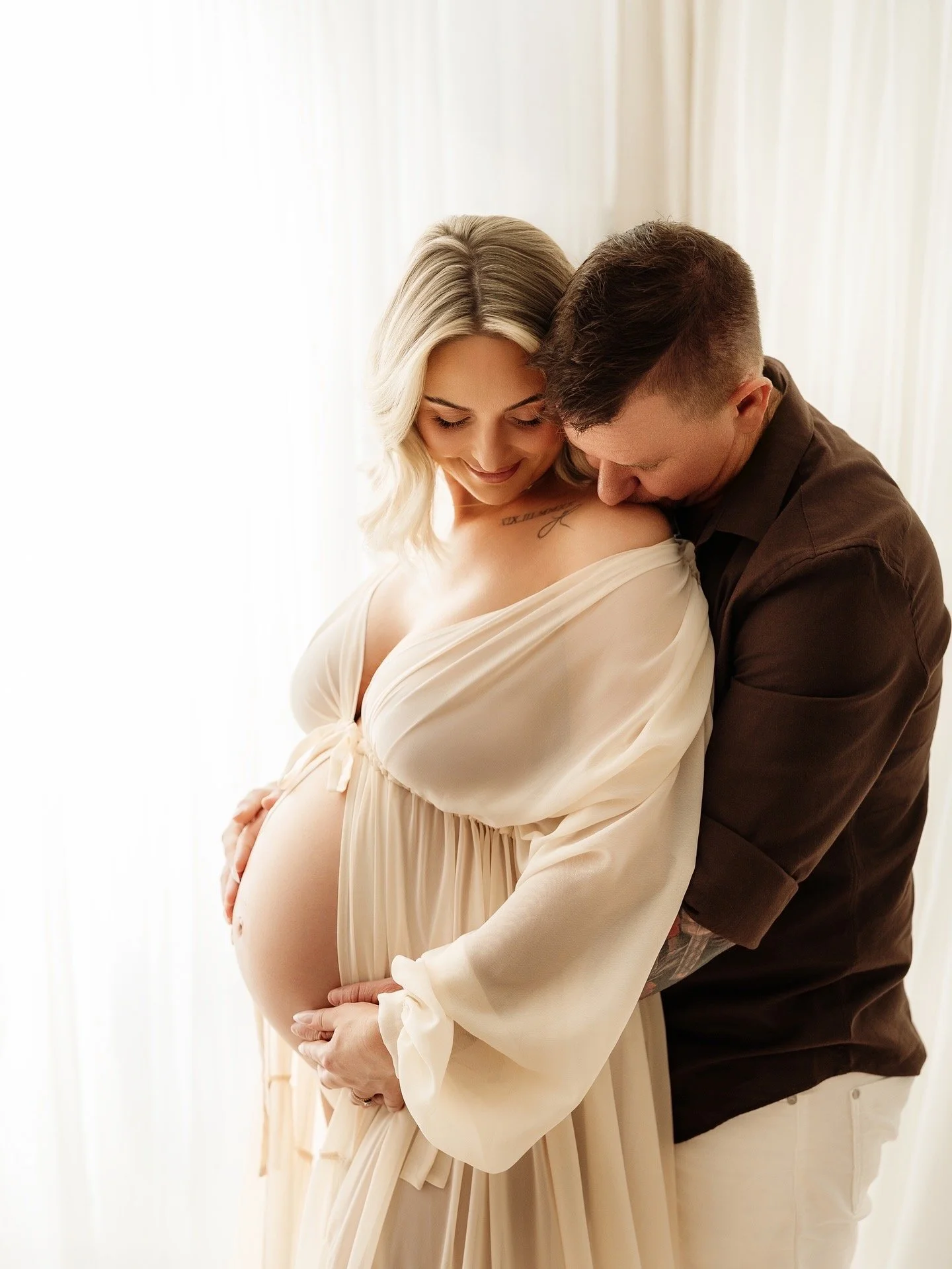 A pregnant woman in a cream dress with a man standing close behind her, both smiling near a bright window.