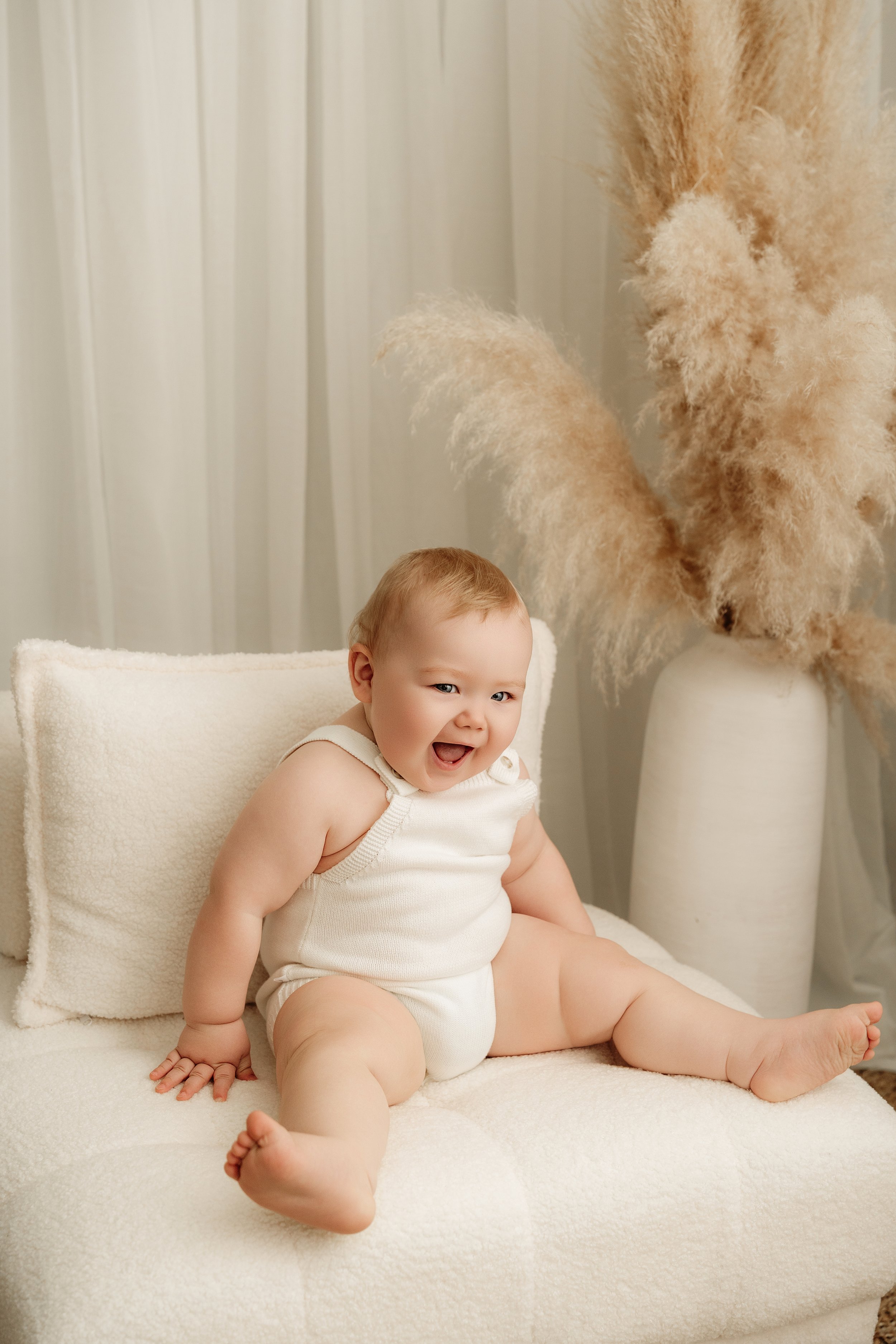 A smiling baby in a white sleeveless romper sits on a white textured chair with a pillow, in front of a vase with tall pampas grass near a sheer curtain.