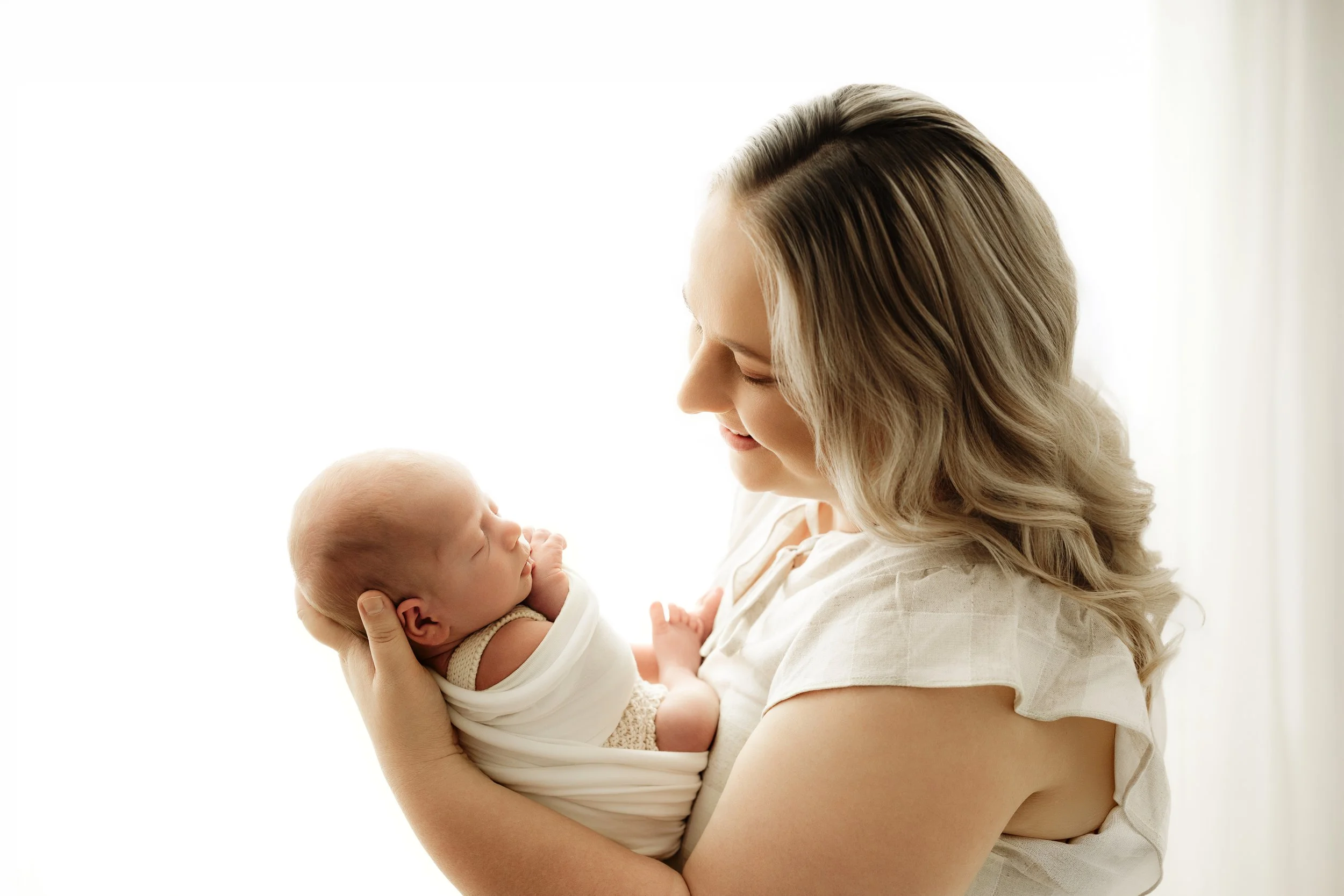 A woman holding a sleeping newborn baby close, with both looking at each other affectionately in a bright, softly-lit room in Brisbane photography studio.