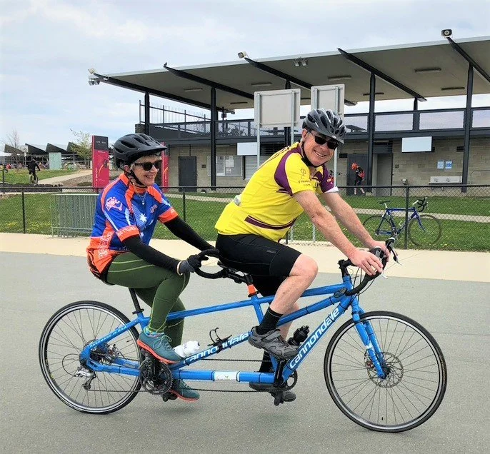 A smiling pilot and stoker riding a blue tandem bicycle together on a Canberra path.