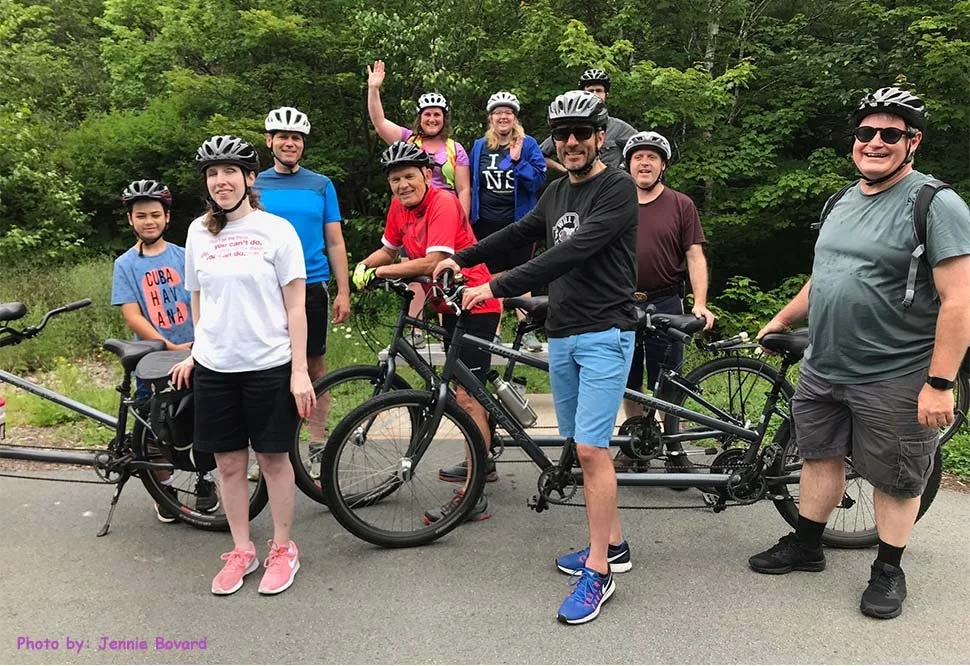 Fitability riders and volunteers standing together with several tandem bicycles on a tree-lined path.