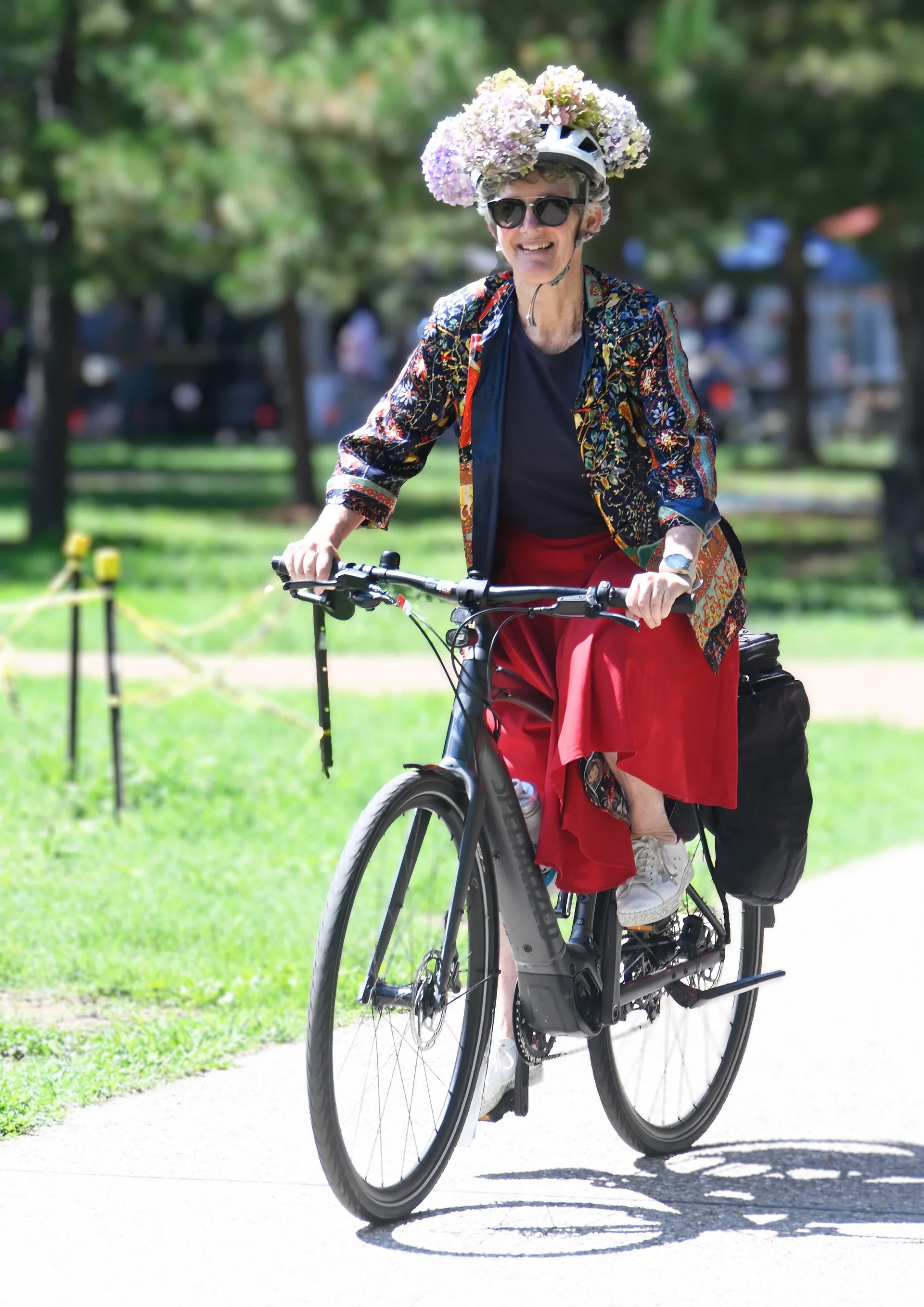 A smiling rider wearing a flower-covered helmet riding an electric bicycle through a Canberra park
