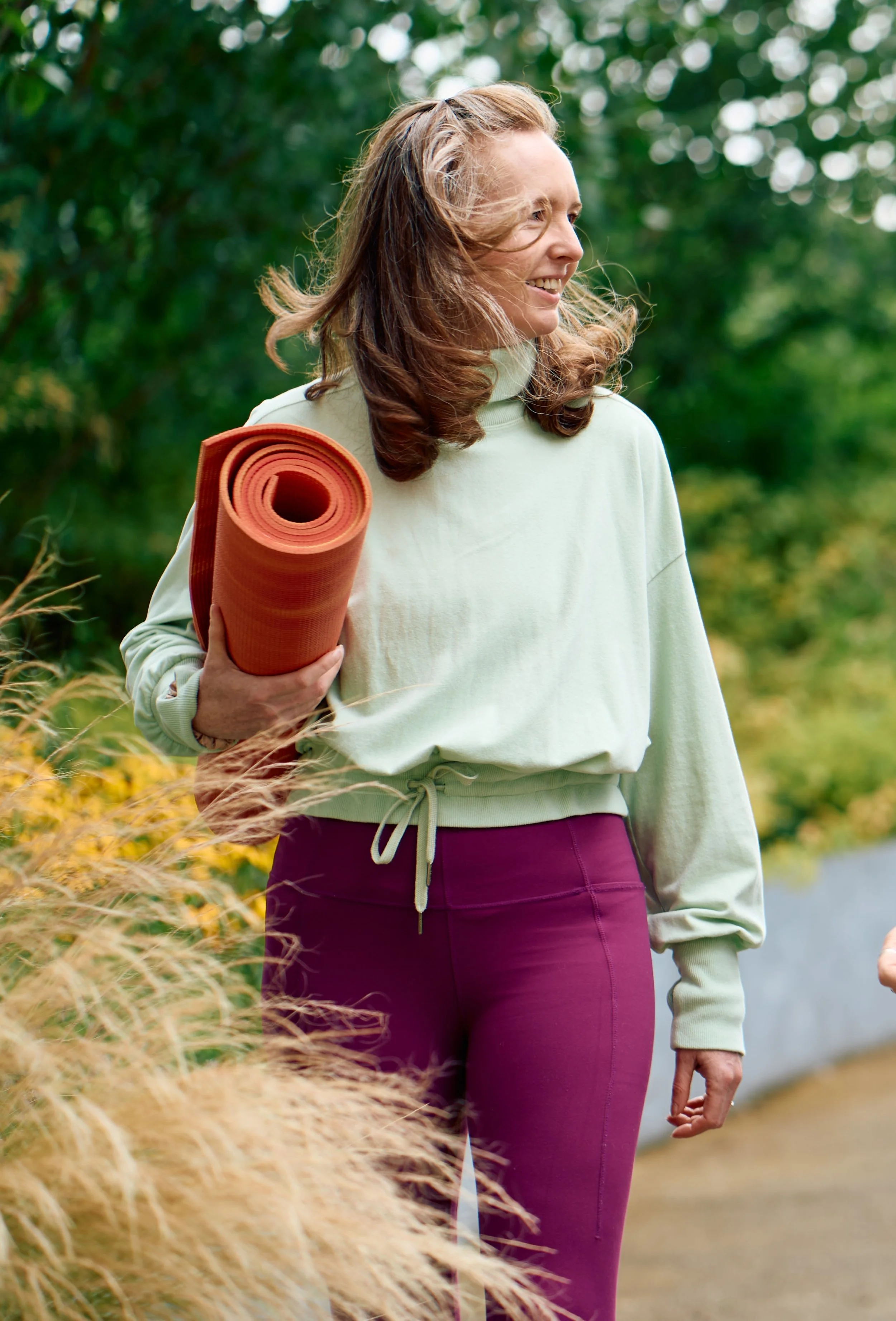 Tula Yoga Jo  holding an orange yoga mat outdoors on a trail surrounded by greenery.