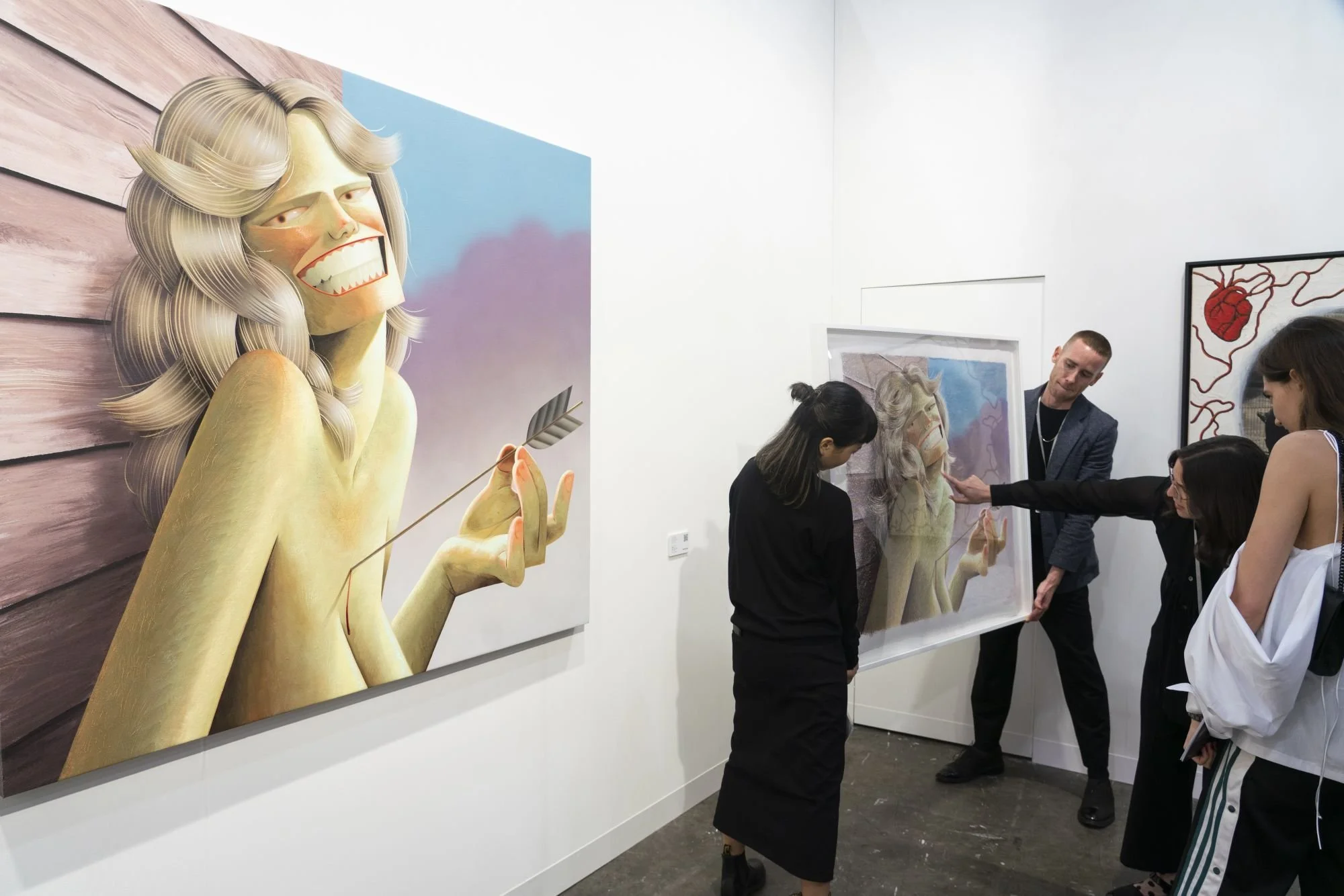 Art gallery scene with a large painting of a figure holding an arrow and smiling. Several people are observing or handling a similar framed artwork. The setting is modern, with plain walls and additional artworks in the background.
