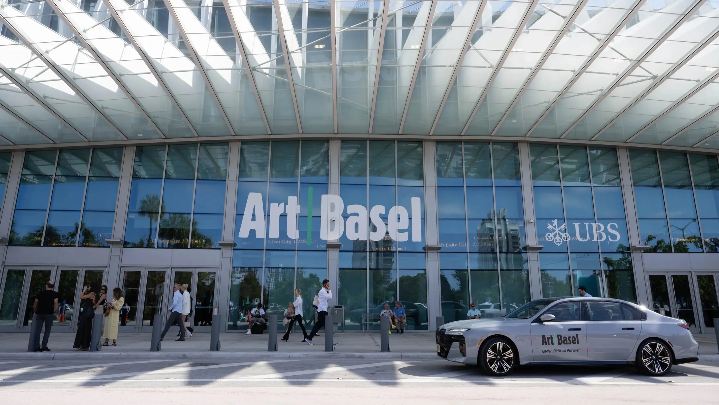 Exterior of Art Basel venue with glass facade, UBS logo, and parked BMW car labeled 'Art Basel.'