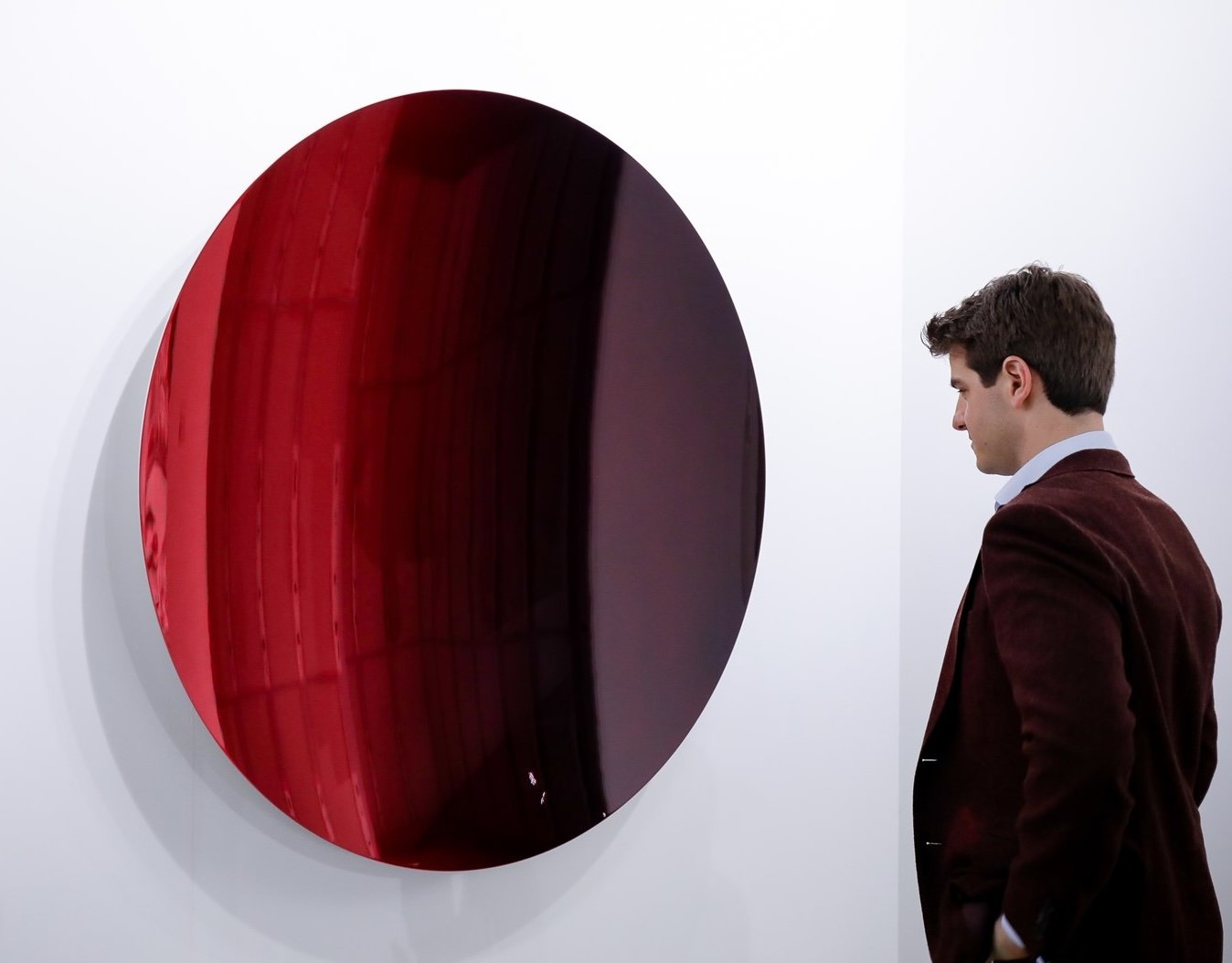 Man observing large reflective red disc artwork on a wall.