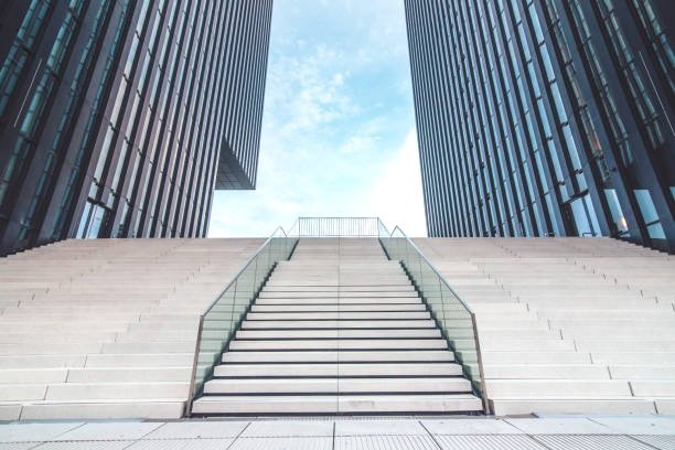 A staircase with glass railings leading up between two tall modern glass office buildings, with a blue sky and some clouds overhead.